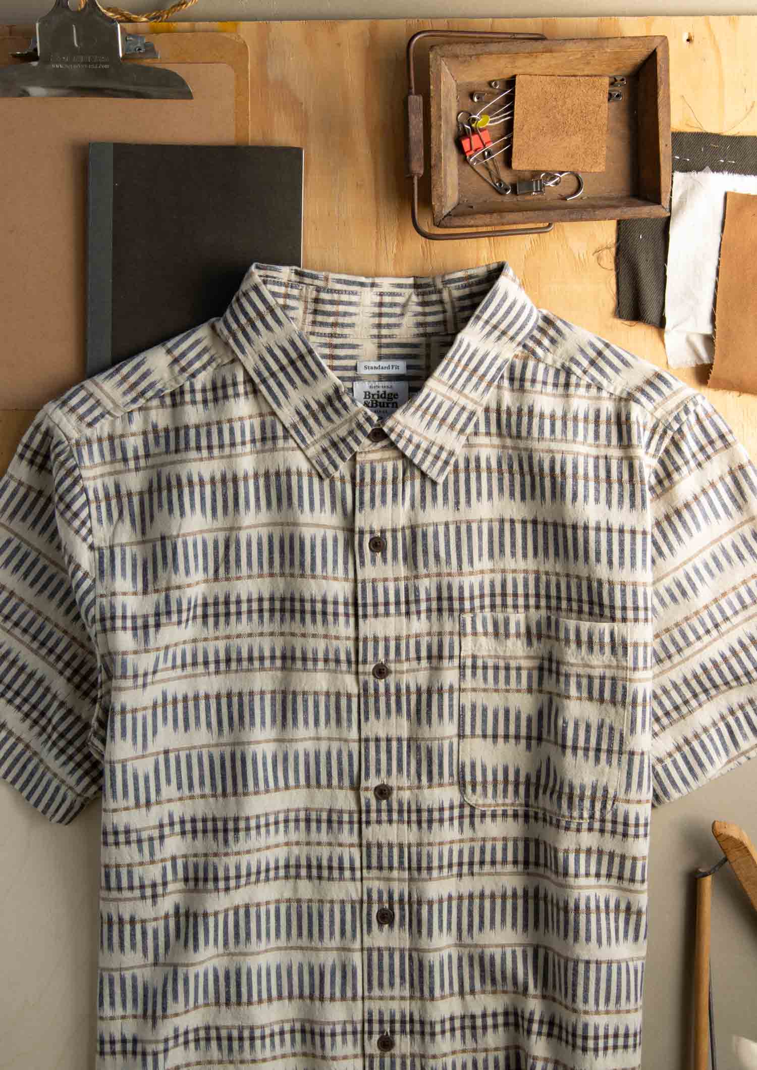 The Bridge & Burn Malcolm Shirt in Mushroom Ikat Stripe—a sustainable, short-sleeve button-up made from soft organic cotton—features a geometric beige, gray, and blue pattern and hangs on a wooden wall among notebooks and tools.