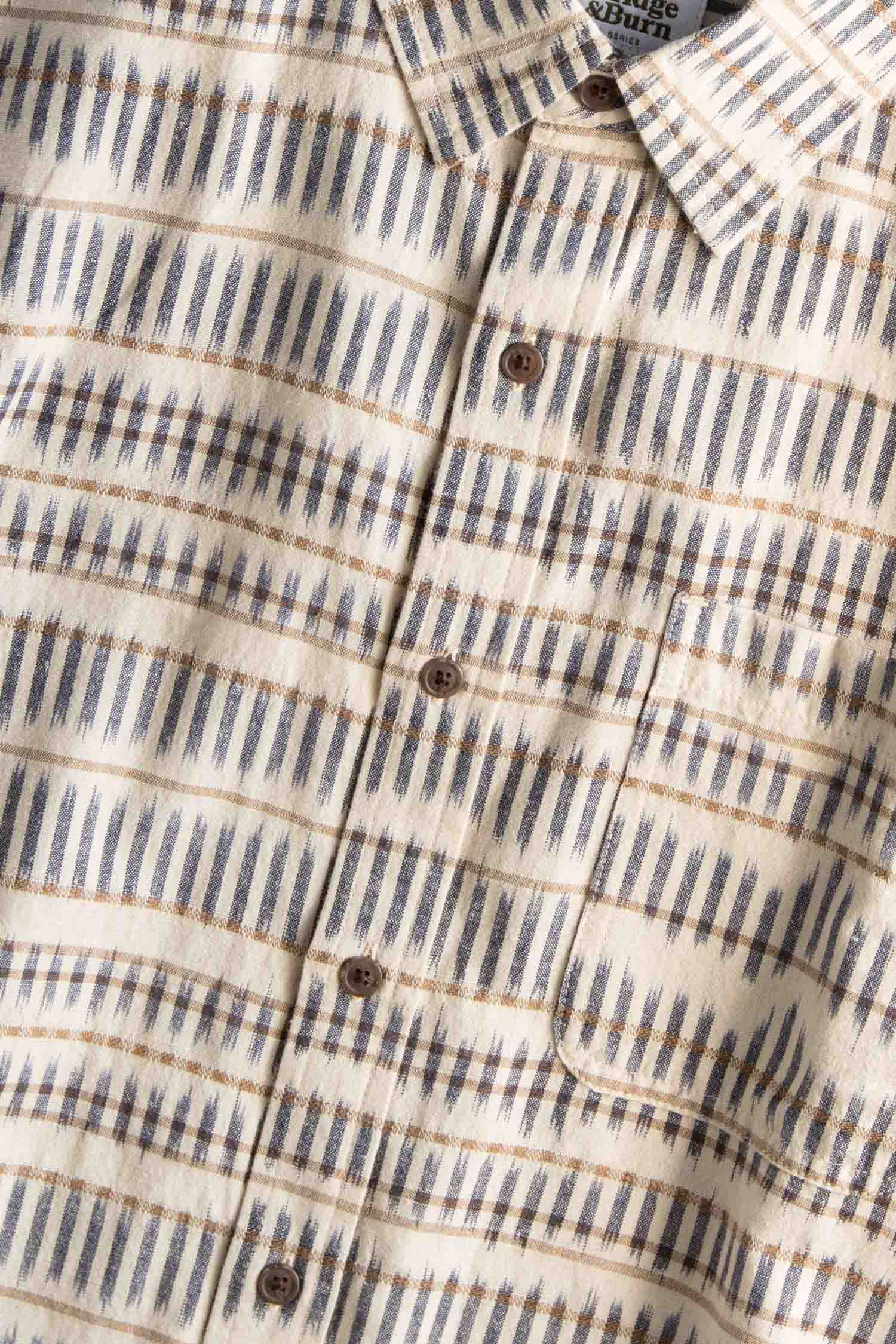The Malcolm Shirt by Bridge & Burn features a cream-colored, short-sleeve button-up design with gray and tan Mushroom Ikat stripes, crafted from organic cotton and finished with brown front buttons.