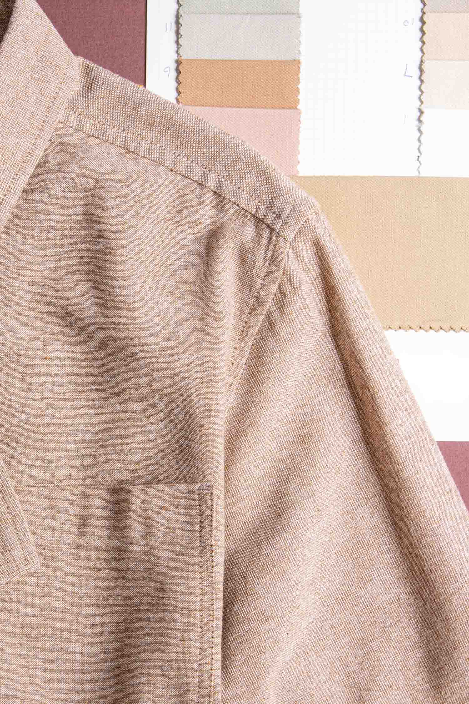 A close-up of the Malcolm Shirt in Rust Chambray by Bridge & Burn shows its shoulder and chest pocket, with neutral-toned fabric swatches in the background, highlighting its eco-friendly design.