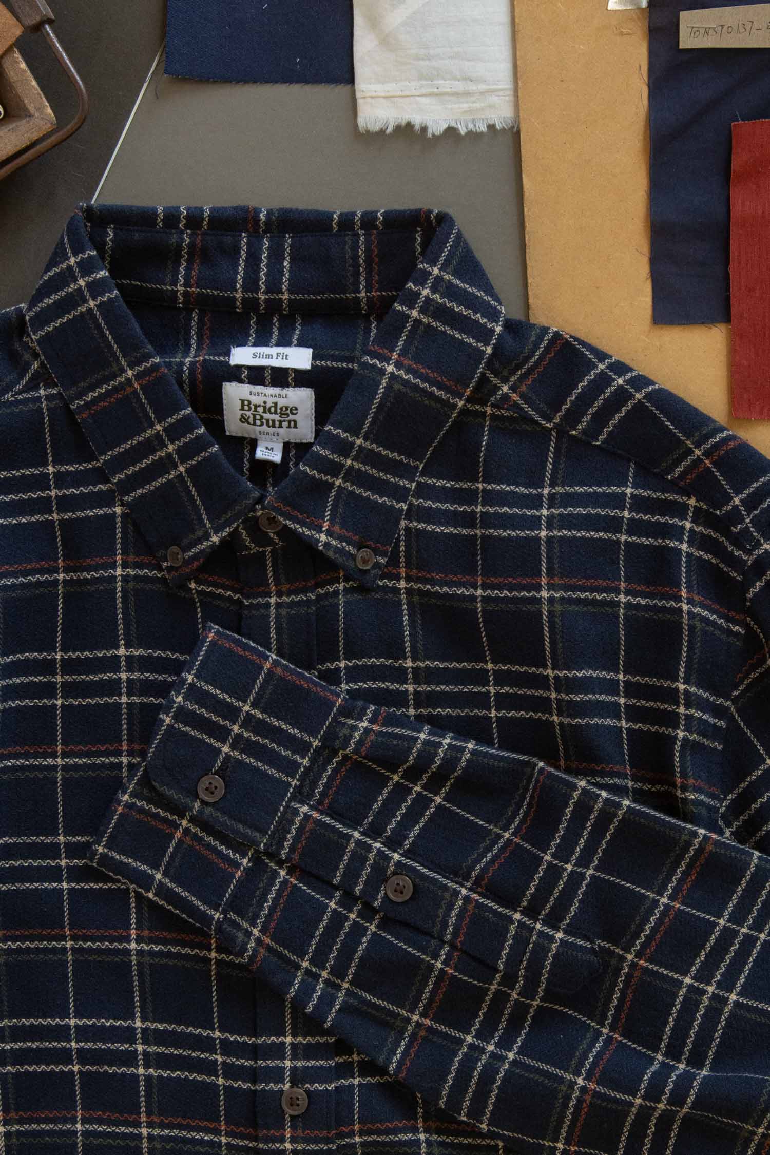 The Bridge & Burn Sutton Slim Shirt in Dusk Herringbone Plaid features a navy blue plaid with beige and red lines, shown flat with fabric swatches and pattern papers, highlighting its minimalist design and refined craftsmanship.