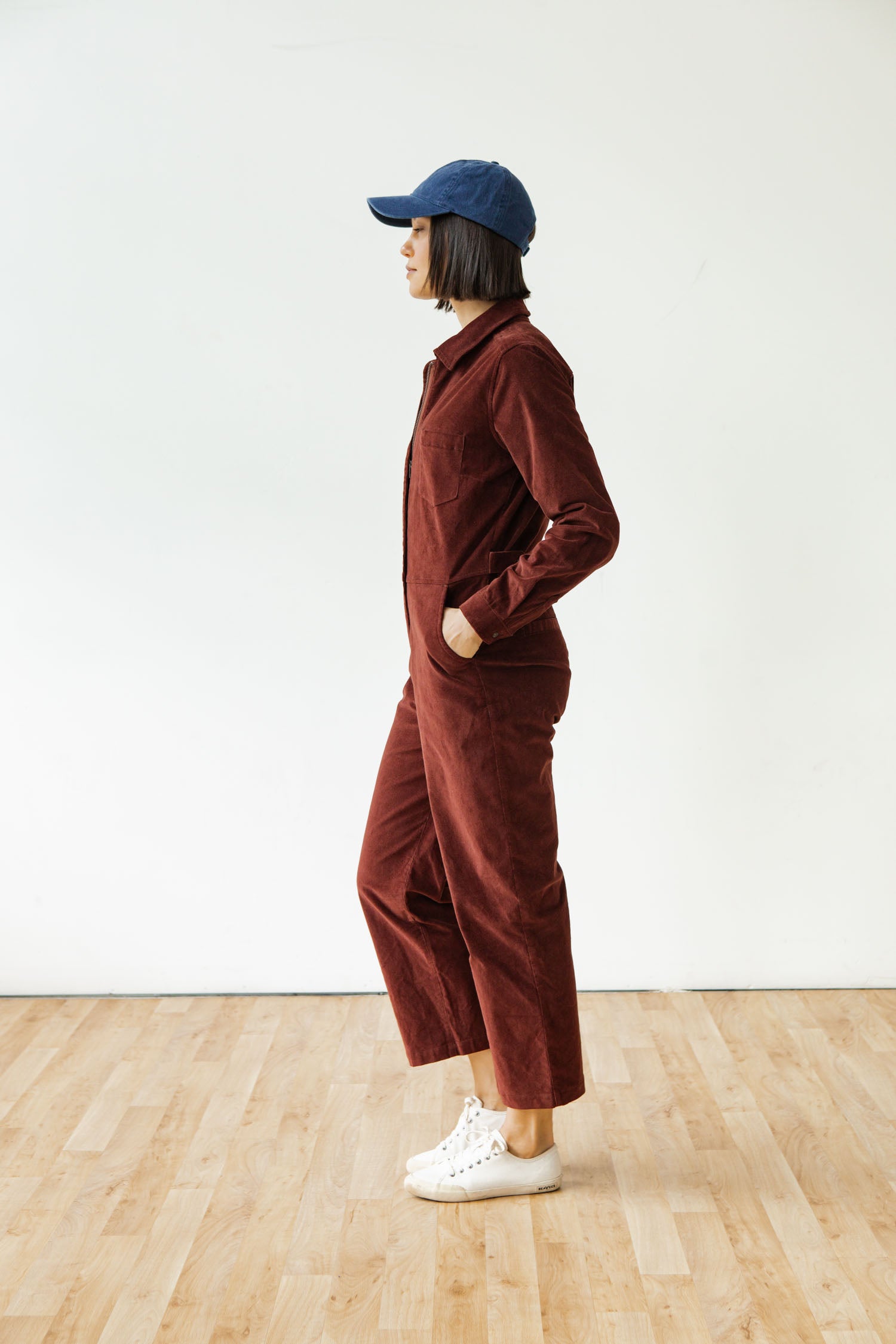 A person wears the Bridge & Burn Faye Jumpsuit in Dark Red Pinwale, paired with white sneakers and a blue cap, standing indoors on a wooden floor against a plain white wall, facing left with hands in pockets.