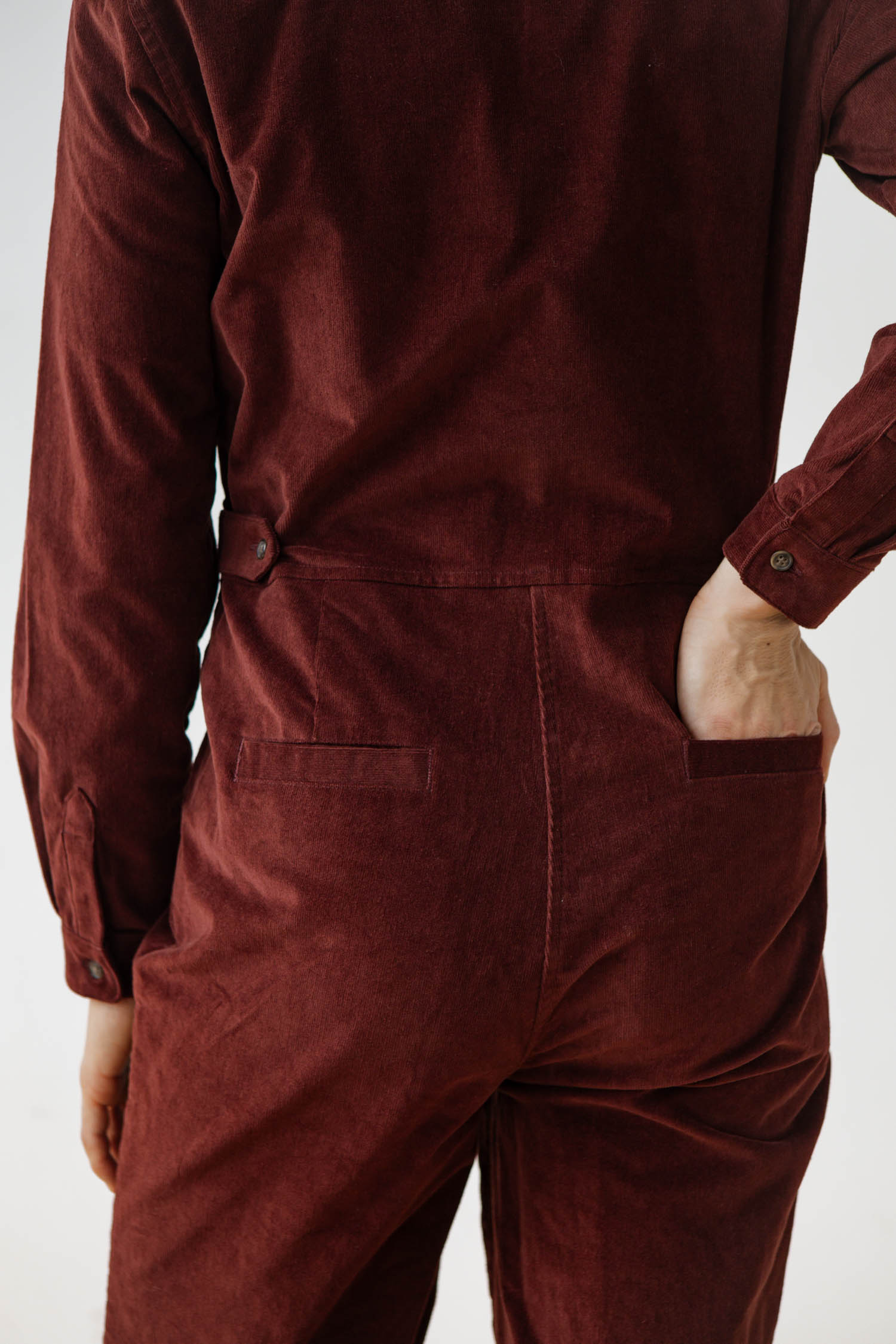 A person stands with their back to the camera, one hand in a pocket, wearing the Bridge & Burn Faye Jumpsuit in Dark Red Pinwale, effortlessly showcasing this stylish women's corduroy workwear piece.