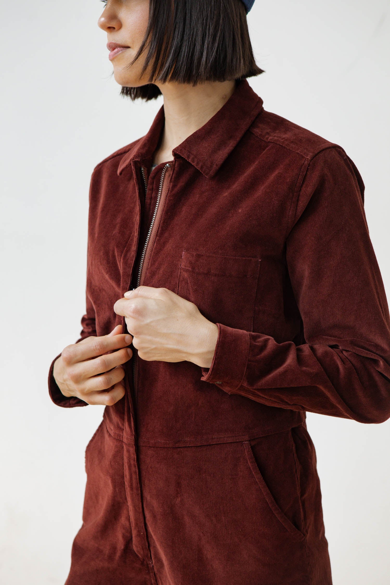 A person with straight, shoulder-length dark hair models the Bridge & Burn Faye Jumpsuit in Dark Red Pinwale, partially unzipped at the front, standing against a plain white background with their face partially out of frame.