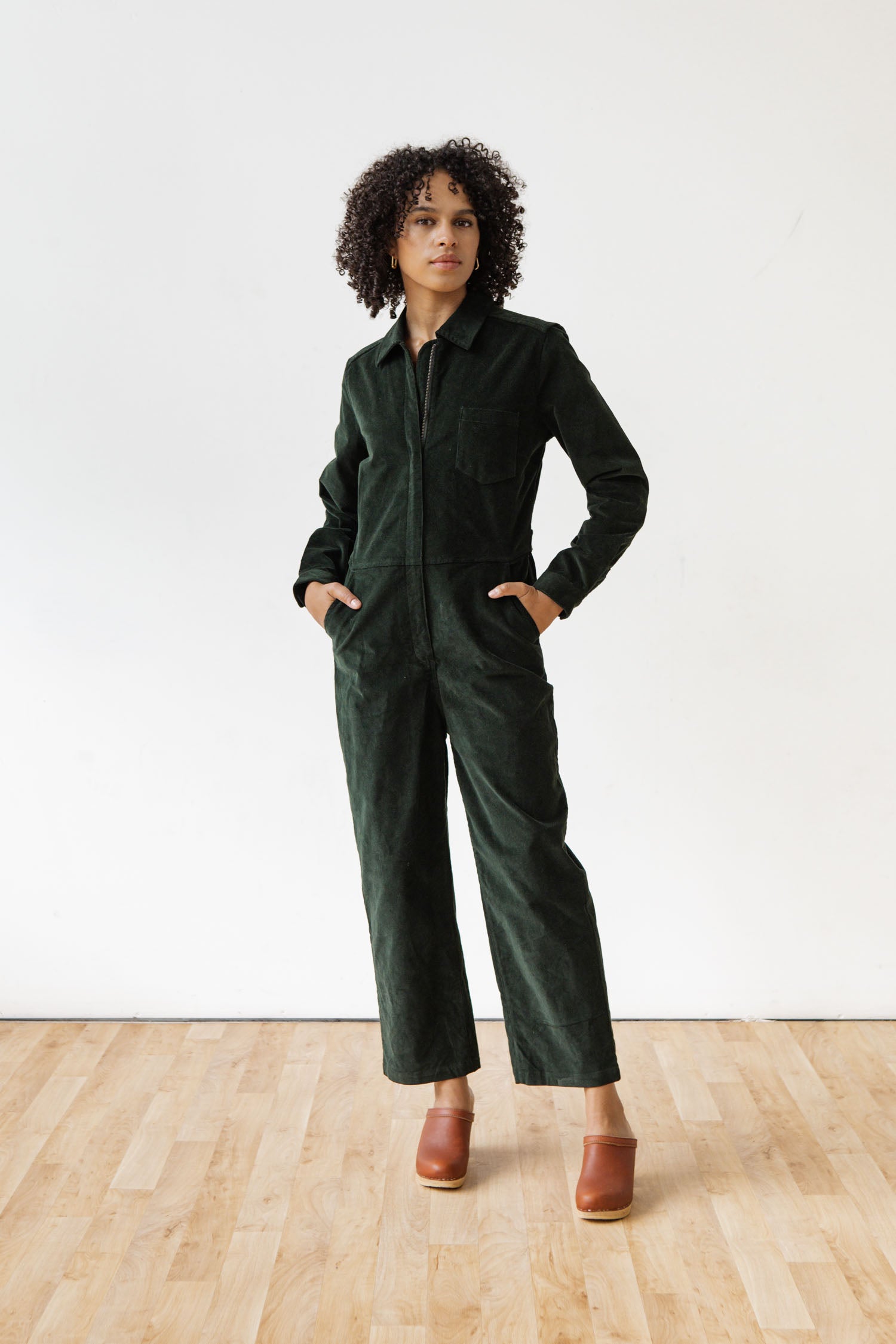 Standing on a wooden floor against a white background, a person with curly hair wears the Bridge & Burn Faye Jumpsuit in Deep Pine Pinwale and brown clogs, hands in pockets, looking to the side.