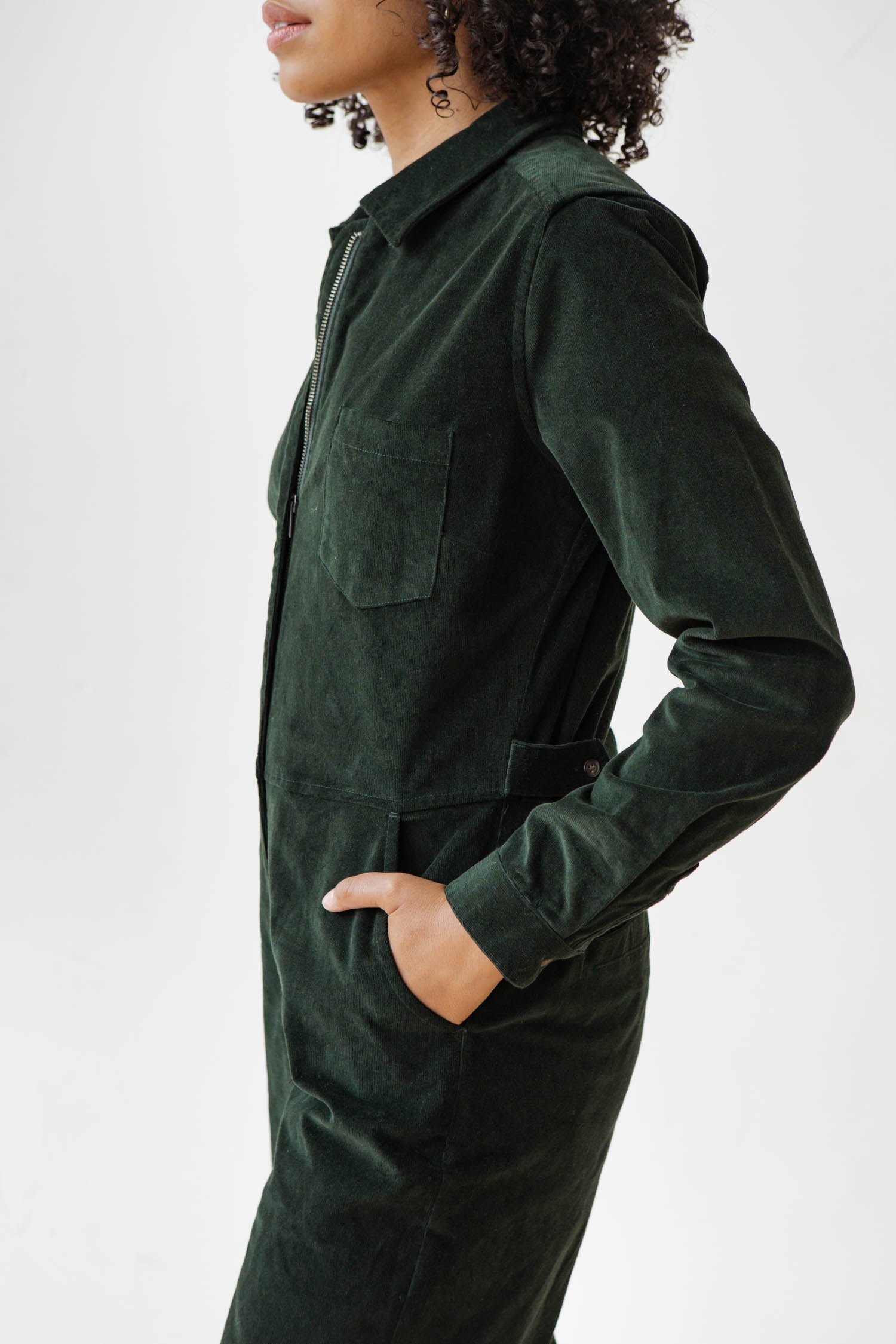 A person with curly hair wears the Bridge & Burn Faye Jumpsuit in Deep Pine Pinwale, hands in side pockets, standing against a plain white background.