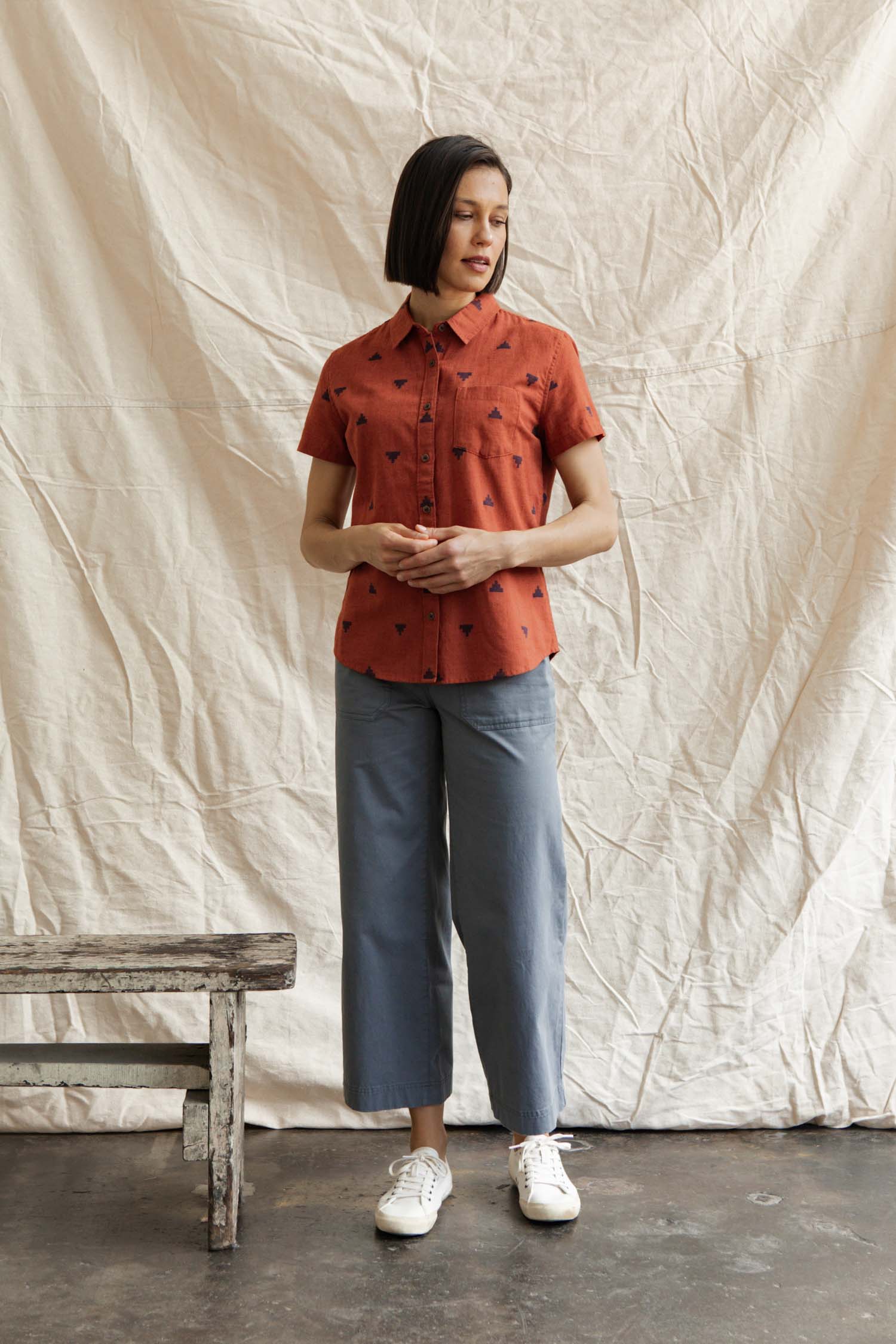 A woman stands indoors before a cream fabric backdrop, wearing the Bridge & Burn Lana Shirt in Rust Fil-Coupé Pyramids, light blue wide-leg pants, and white sneakers. A rustic wooden bench is to her left.