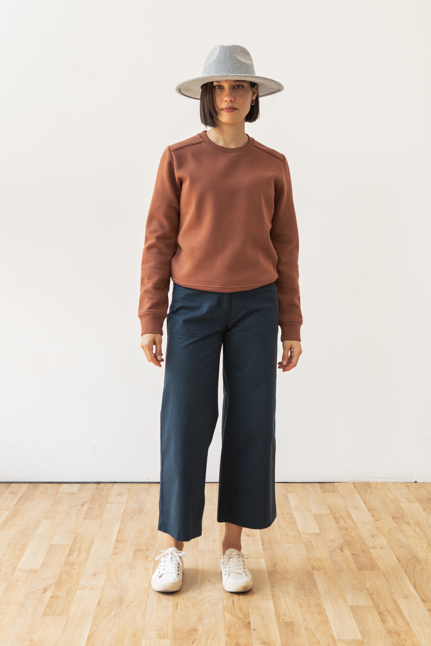 A woman stands indoors on a wooden floor, wearing Bridge & Burn's Bailey Crew Sweatshirt in Burnt Umber, a gray wide-brim hat, blue wide-leg pants, and white sneakers against a plain white wall.