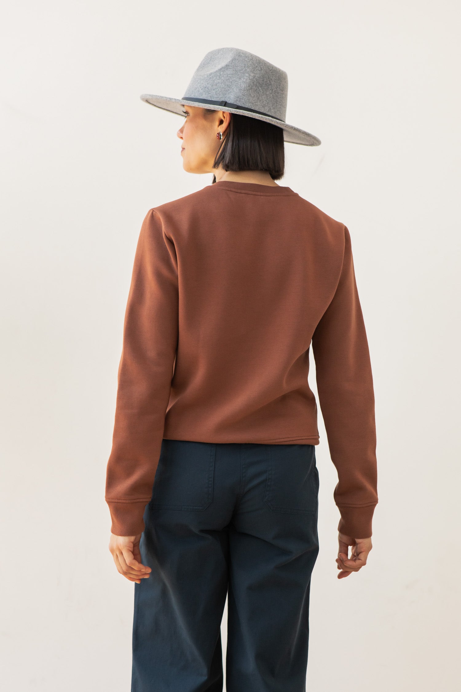 A person wearing the Bridge & Burn Bailey Crew Sweatshirt in Burnt Umber, a gray wide-brimmed hat, and dark pants stands facing away from the camera against a plain light background.
