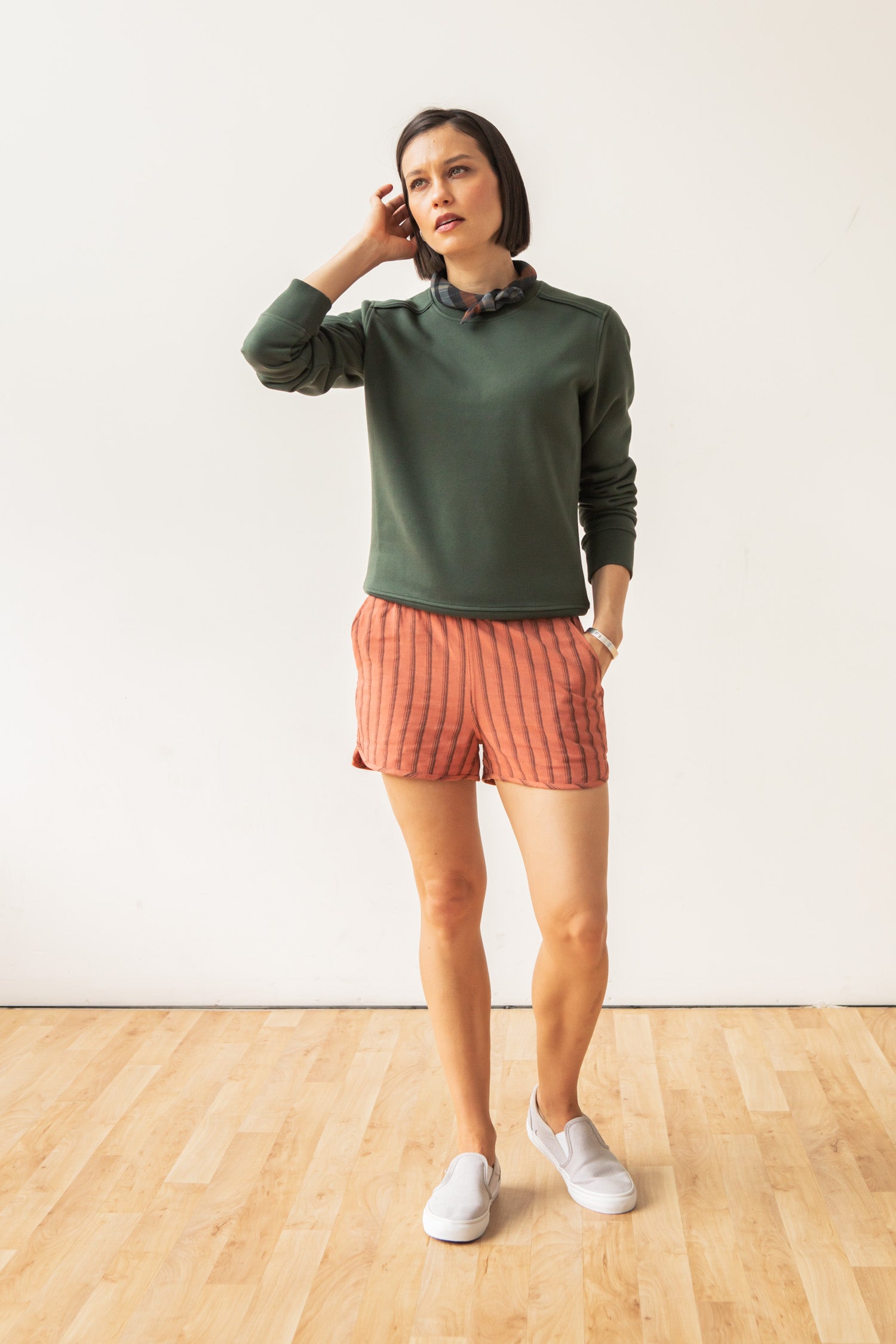 A woman stands indoors on a wooden floor, wearing the Bailey Crew Sweatshirt in Forest Night by Bridge & Burn, coral striped shorts, white slip-on shoes, and a dark scrunchie. One hand touches her head as she maintains a neutral expression.