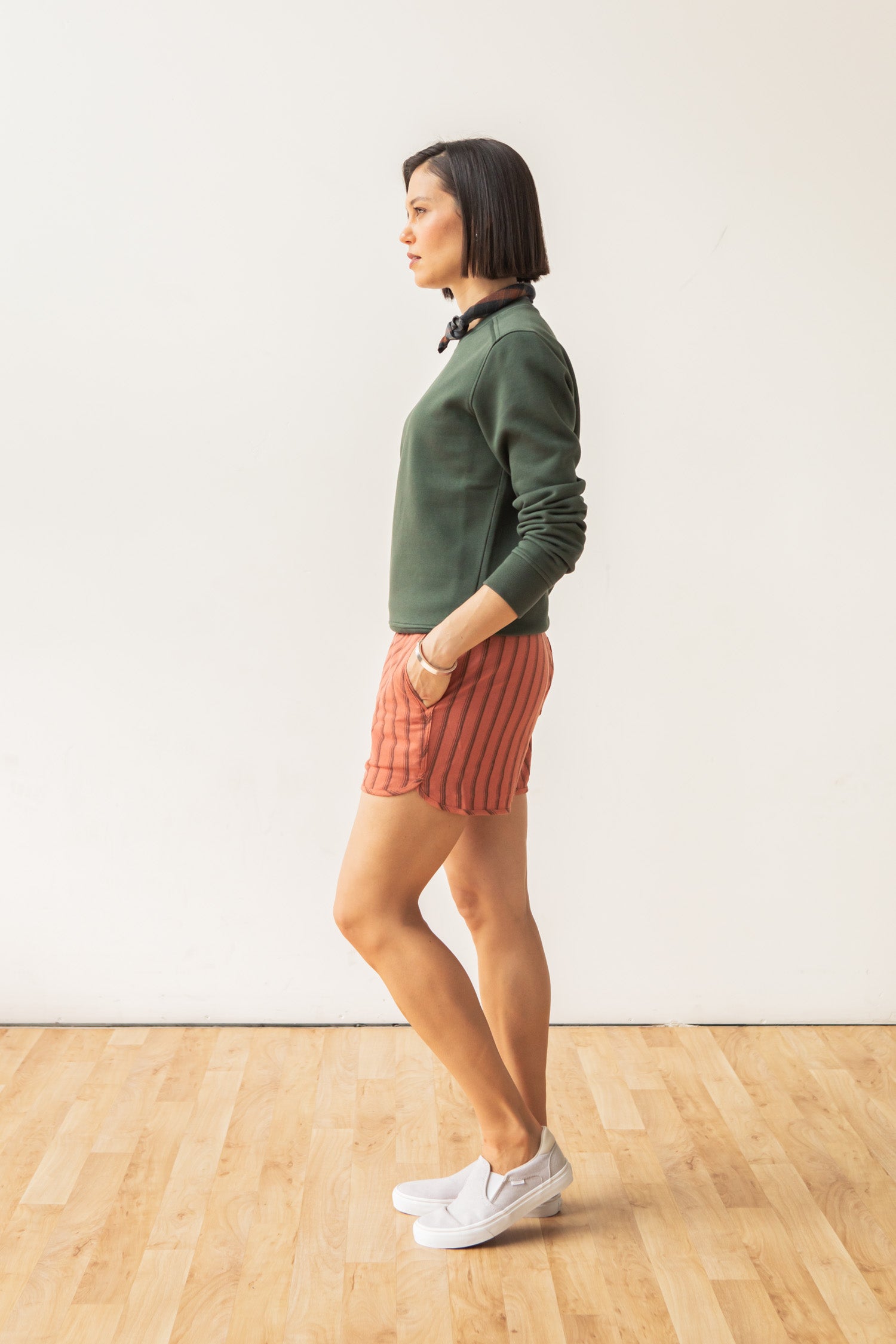 A woman with short dark hair stands in profile against a white wall, wearing the Bridge & Burn Bailey Crew Sweatshirt in Forest Night, red striped shorts, and white sneakers, hands in pockets on a wooden floor.