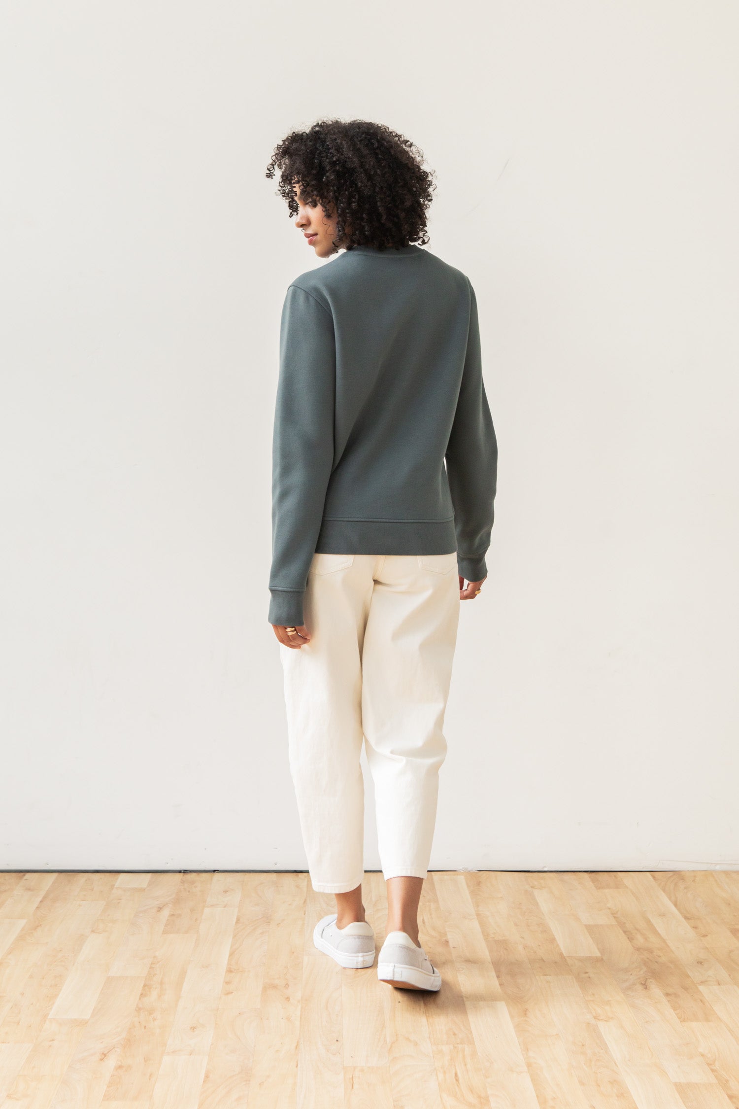 A person with curly hair, dressed in the Bridge & Burn Bailey Crew Sweatshirt in Slate, white pants, and gray slip-on shoes, stands on a wooden floor facing away from the camera against a plain white wall.