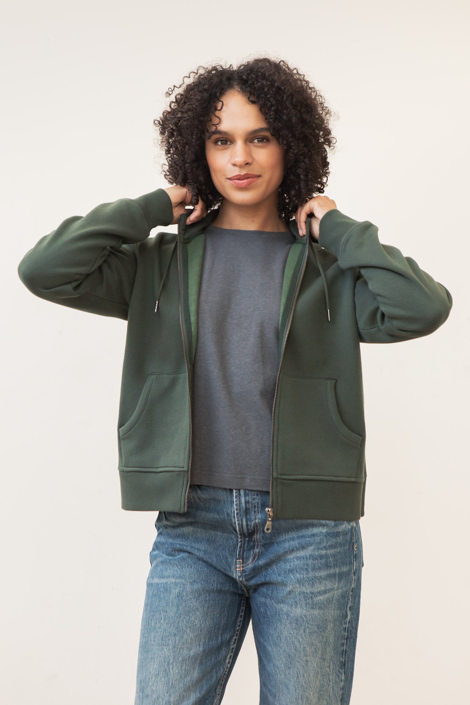 A person with curly hair wears the Bridge & Burn Mason Hoodie in Forest Night over a gray shirt and blue jeans, holding the hoodie by the collar and facing the camera against a plain light background.