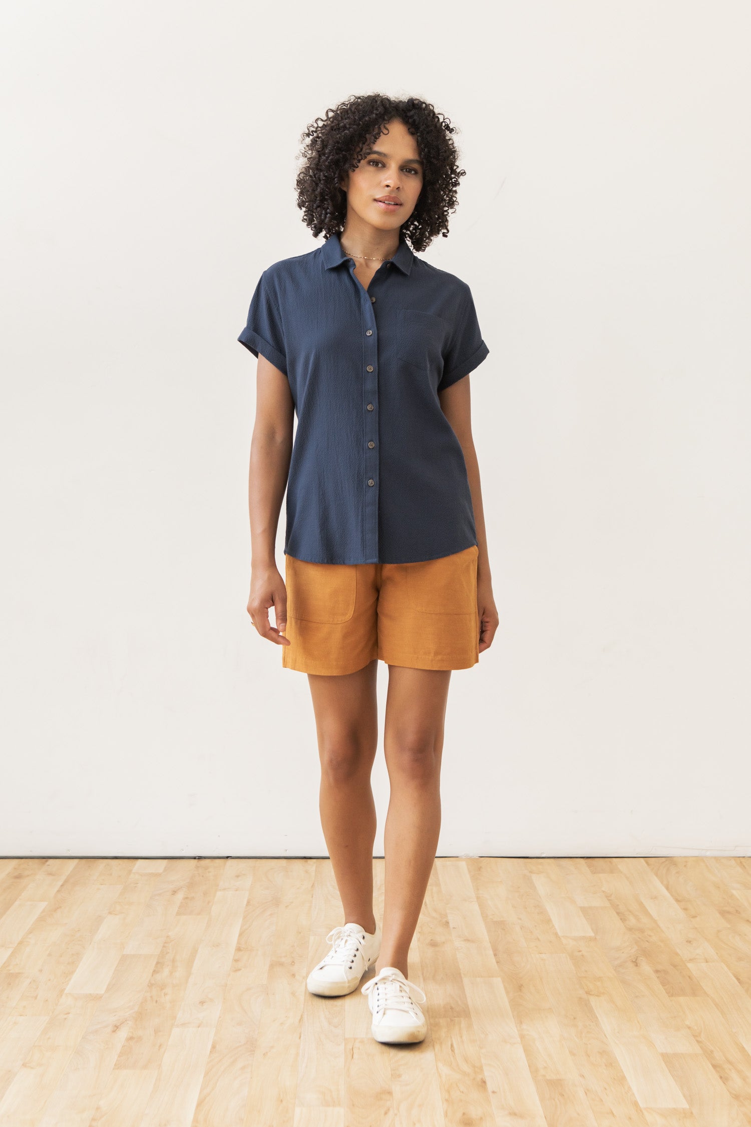 A woman with curly hair stands indoors on a wooden floor, wearing the Bridge & Burn Bea Shirt in Navy Seersucker, mustard-colored shorts, and white sneakers against a plain white wall.