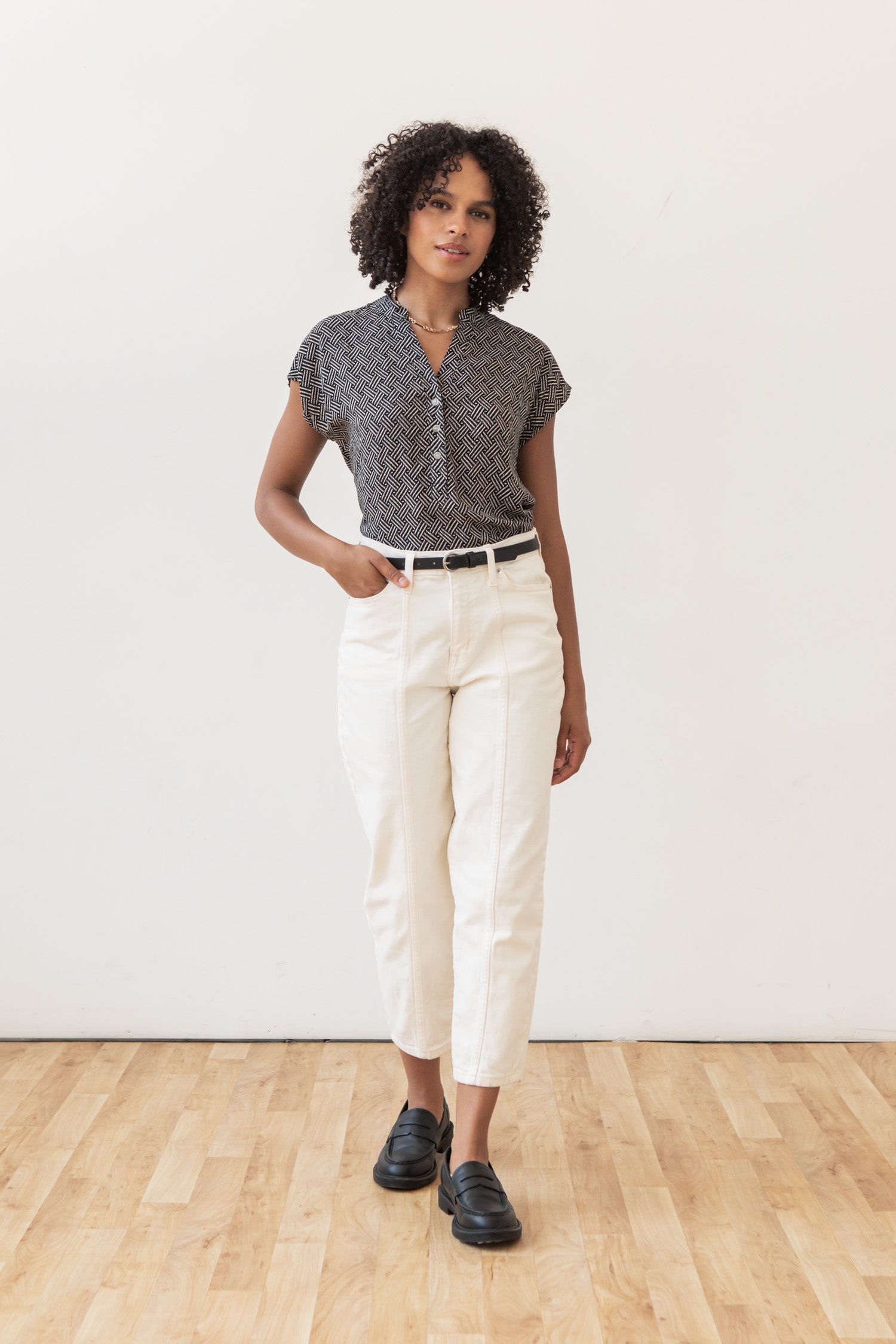A woman with curly hair stands on a wooden floor, wearing the Bridge & Burn Ladd Shirt in Black Geo, white high-waisted pants with a black belt, and black loafers. One hand is in her pocket as she looks forward.