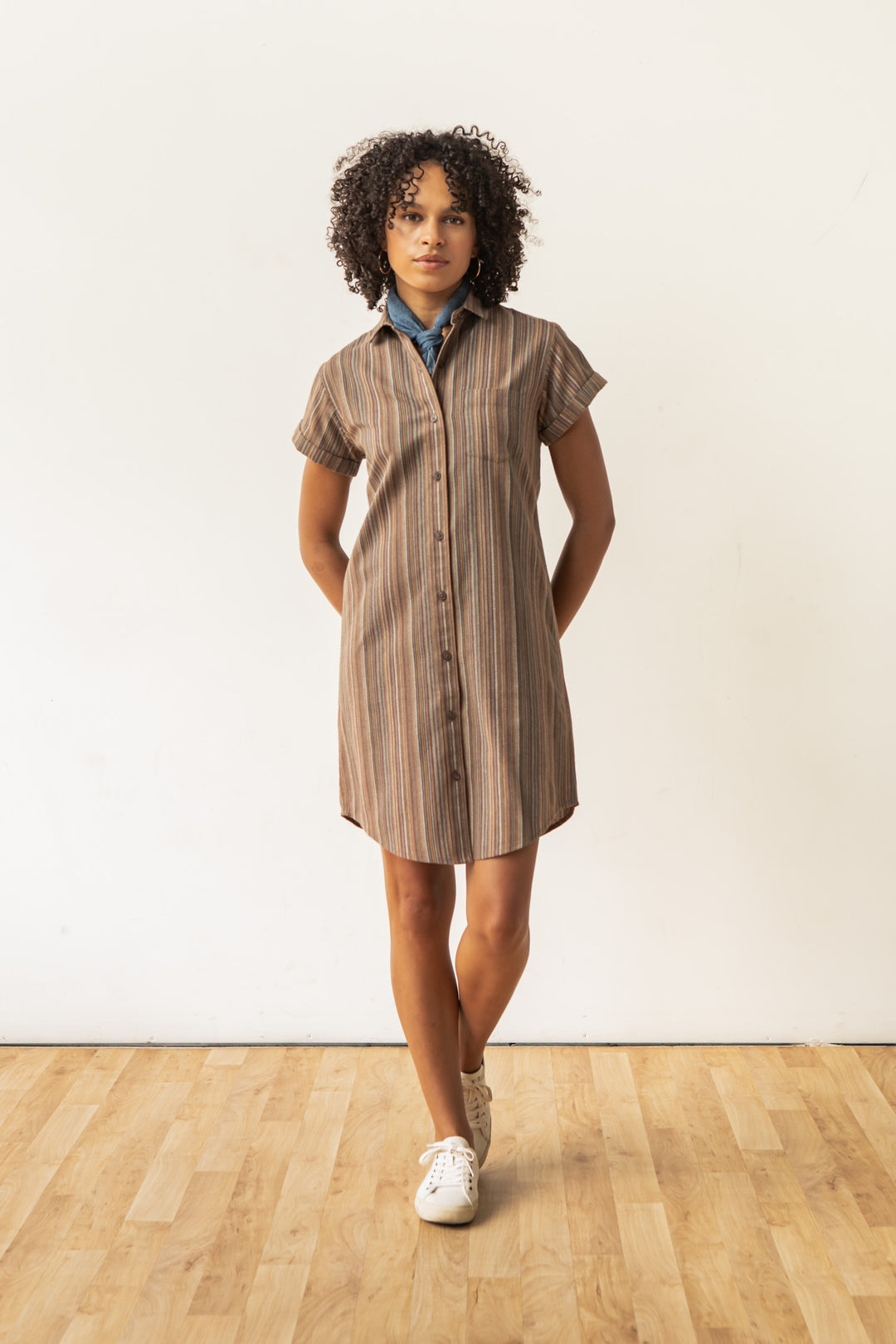 A woman with curly hair stands indoors on a wooden floor, facing the camera with her hands behind her back. She wears the Bridge & Burn Loren Shirt Dress in Multistripe, set against a plain white wall.