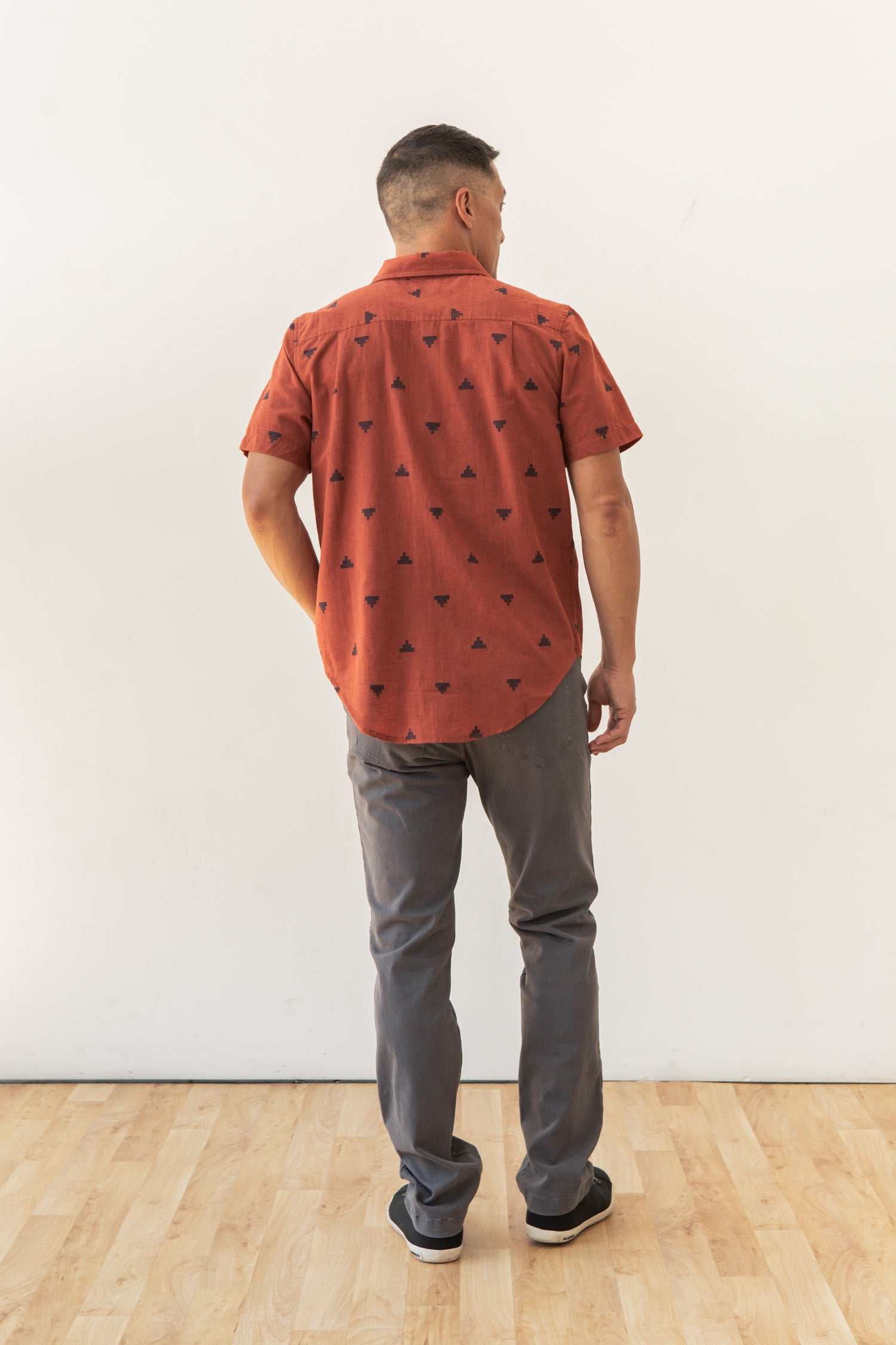 A man stands indoors on a wooden floor facing away, wearing the Bridge & Burn Malcolm Shirt in Rust Fil-Coupé Pyramids, paired with gray pants and black shoes. The background is a plain white wall.