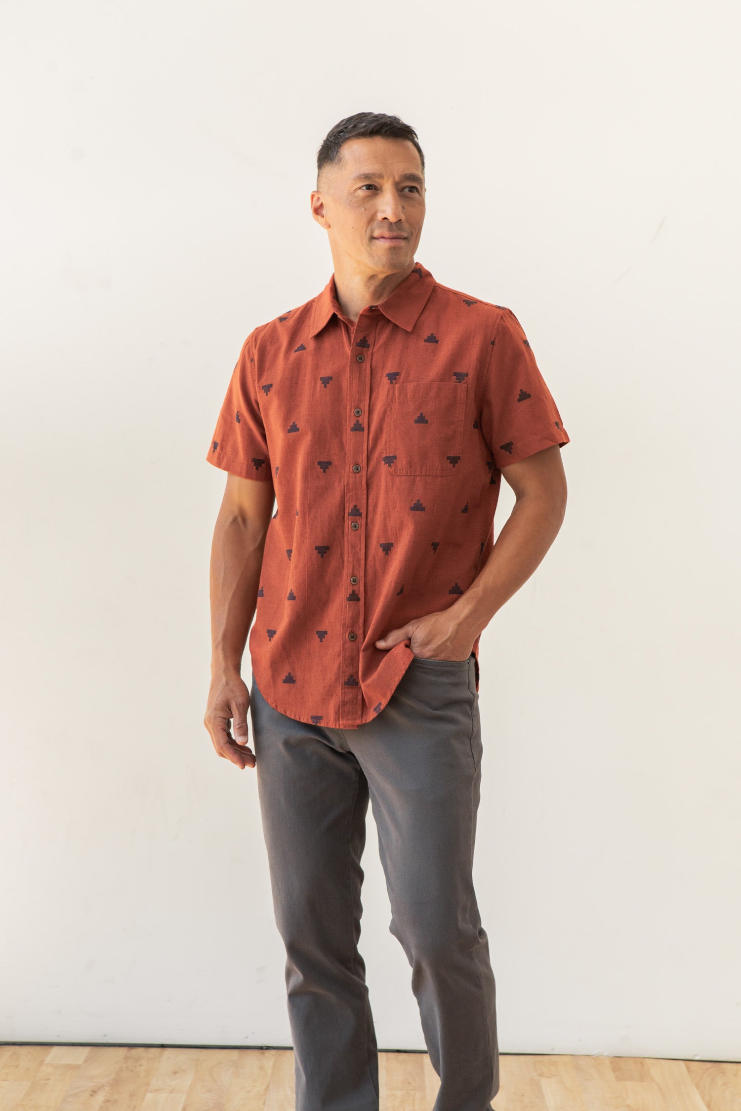 A man stands indoors against a plain light wall, wearing Bridge & Burn's Malcolm Shirt in Rust Fil-Coupé Pyramids—a hemp short sleeve button-up with a subtle black pattern—and gray pants, with one hand in his pocket.