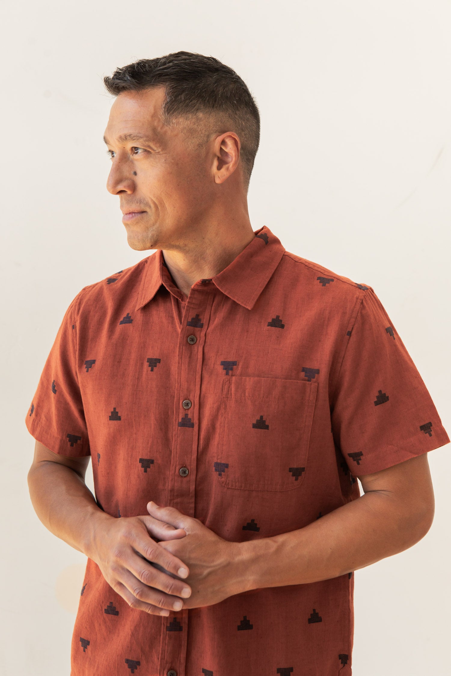 A man with short dark hair wears the Bridge & Burn Malcolm Shirt in Rust Fil-Coupé Pyramids—a sustainable button-up with a subtle black geometric pattern—as he stands against a plain light background, looking off to the side with hands loosely clasped.