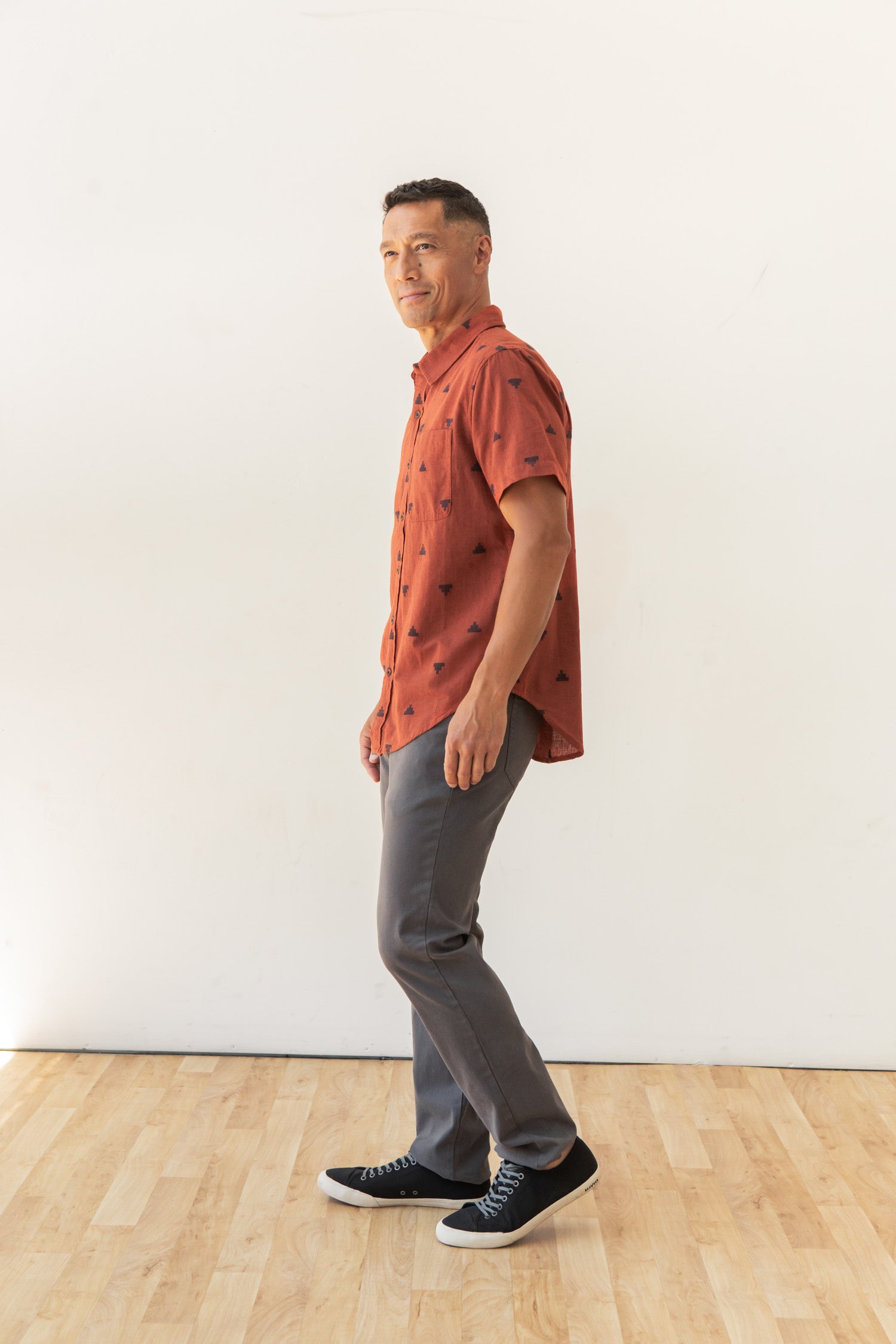 A man stands sideways on a wooden floor against a plain white wall, wearing the Bridge & Burn Malcolm Shirt in Rust Fil-Coupé Pyramids, paired with gray pants and black sneakers.