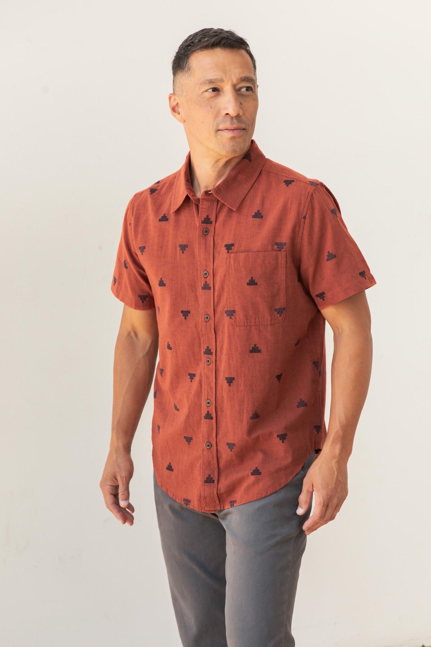 A man wears the Bridge & Burn Malcolm Shirt in Rust Fil-Coupé Pyramids with gray pants, standing against a plain light background and looking off to the side.