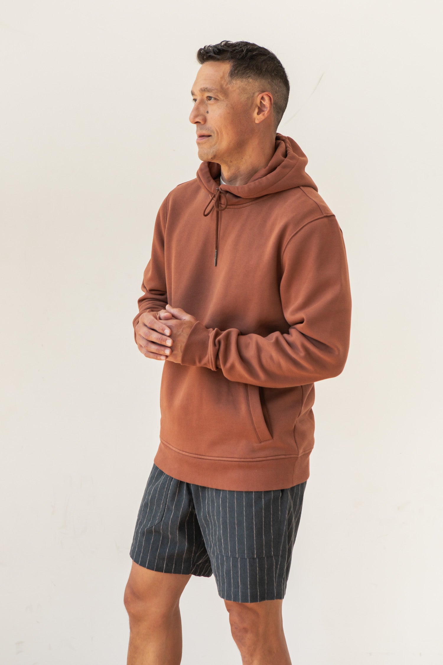 A man wearing the Bridge & Burn Williams Pullover Hoodie in Burnt Umber and pinstriped shorts stands against a plain white background, looking slightly to the side with his hands clasped together.