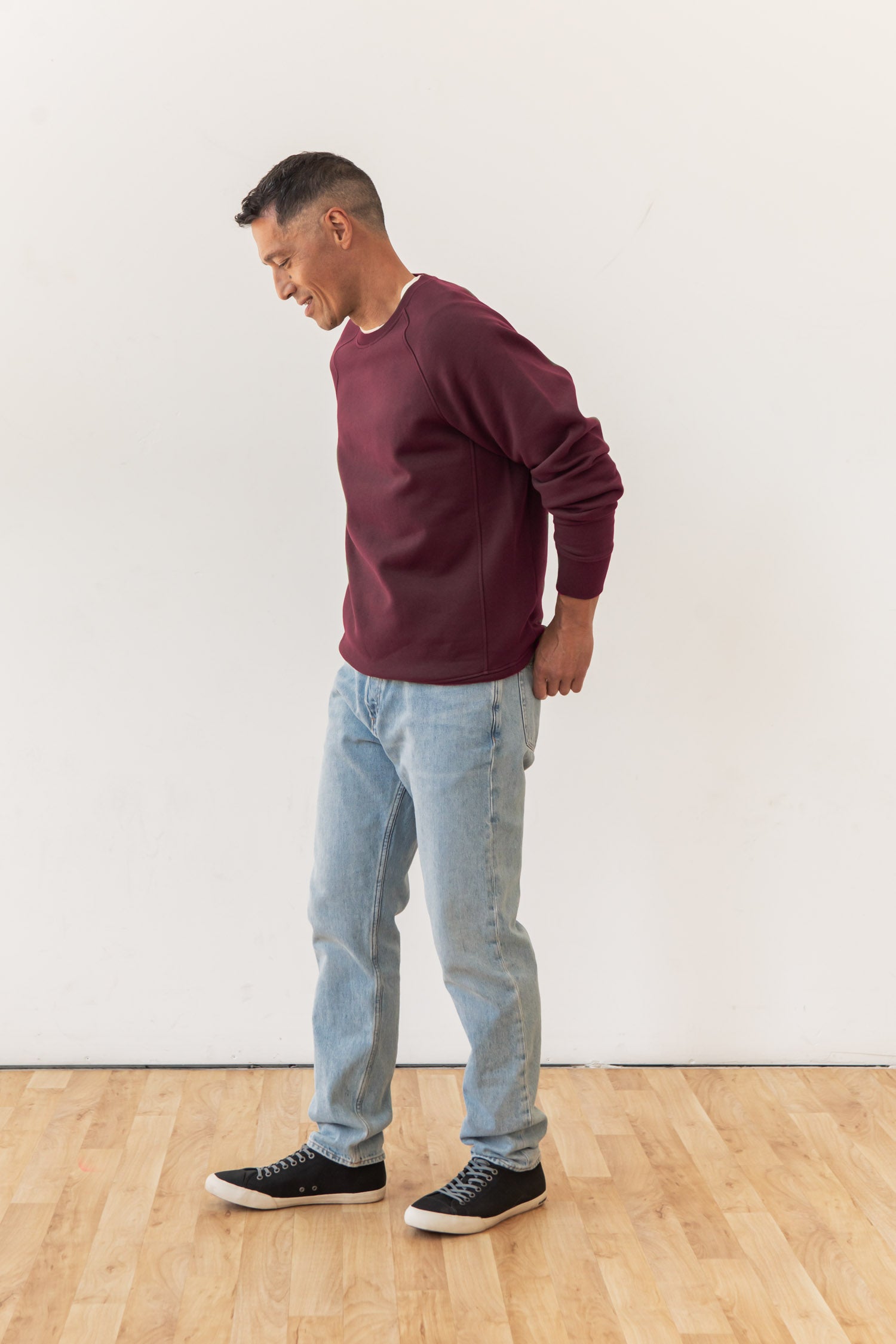 A man wearing a Bridge & Burn Lincoln Crew Sweatshirt in burgundy, light blue jeans, and black sneakers stands on a wooden floor against a plain white wall, looking down and smiling.