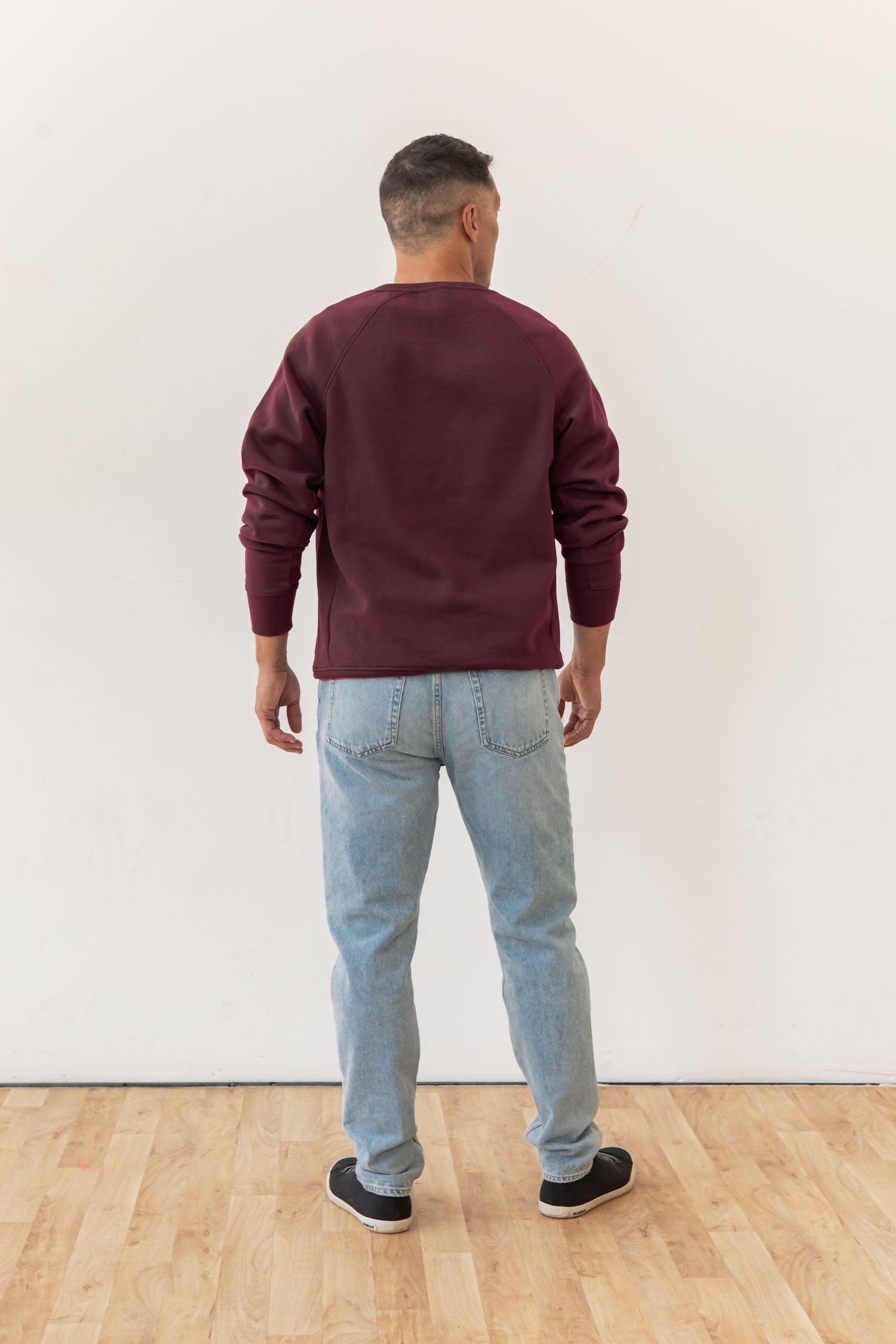 A man stands with his back to the camera, wearing the Bridge & Burn Lincoln Crew Sweatshirt in Burgundy, light blue jeans, and black sneakers on a light wooden floor against a plain white wall.