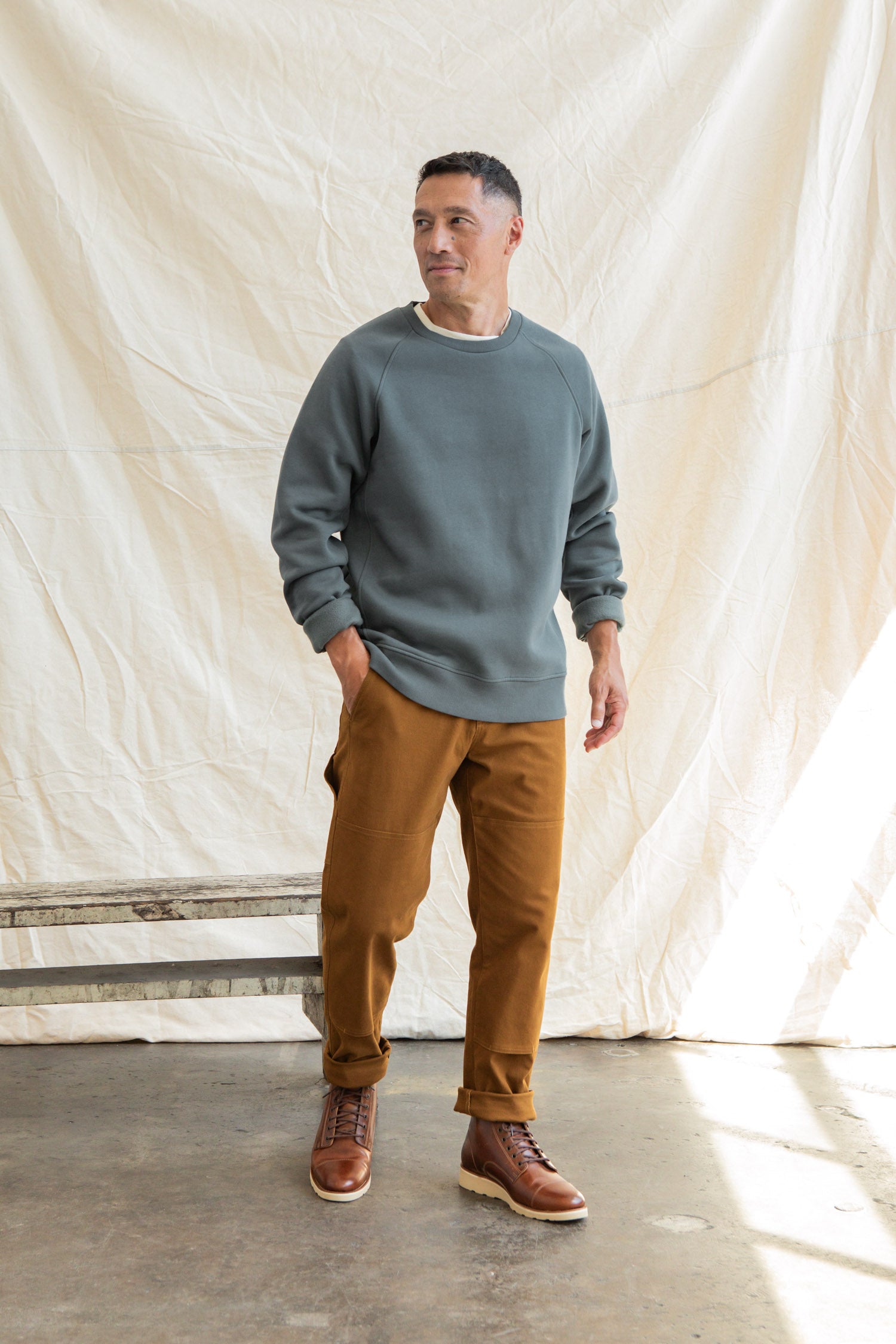 A man stands indoors on a concrete floor, wearing the Bridge & Burn Lincoln Crew Sweatshirt in Slate, brown pants, and brown leather boots. A light-colored fabric backdrop is behind him as he looks slightly to the side with a relaxed posture.