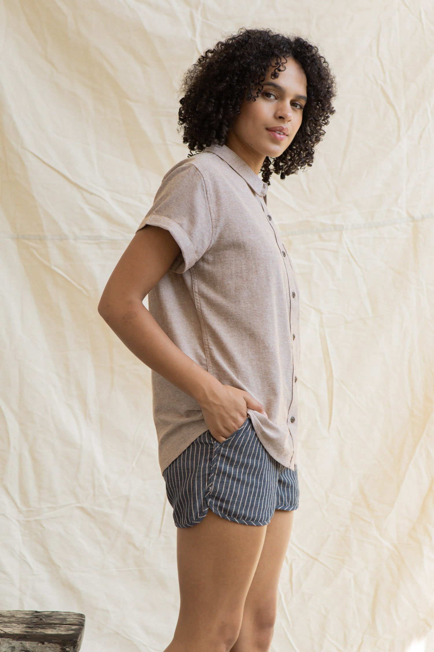 A person with curly hair stands sideways, facing the camera, wearing the Bridge & Burn Bea Shirt in Rust Chambray and blue pinstriped shorts. One hand is in their pocket as they pose against a light fabric background.
