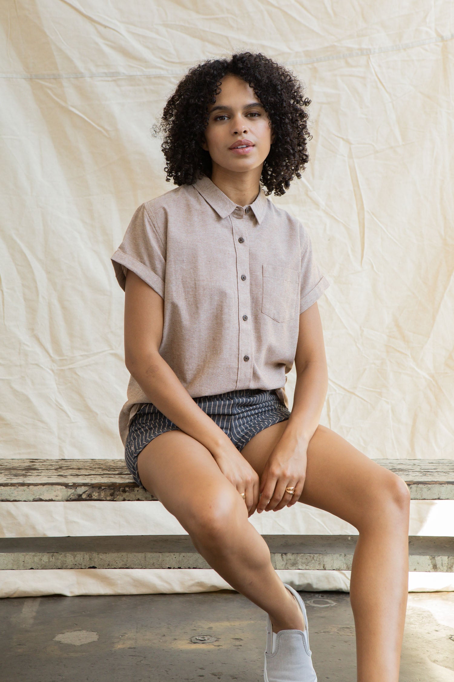 A woman with curly hair wears the Bridge & Burn Bea Shirt in Rust Chambray and patterned shorts, sitting on a wooden bench against a light fabric backdrop. She looks at the camera with a relaxed expression.