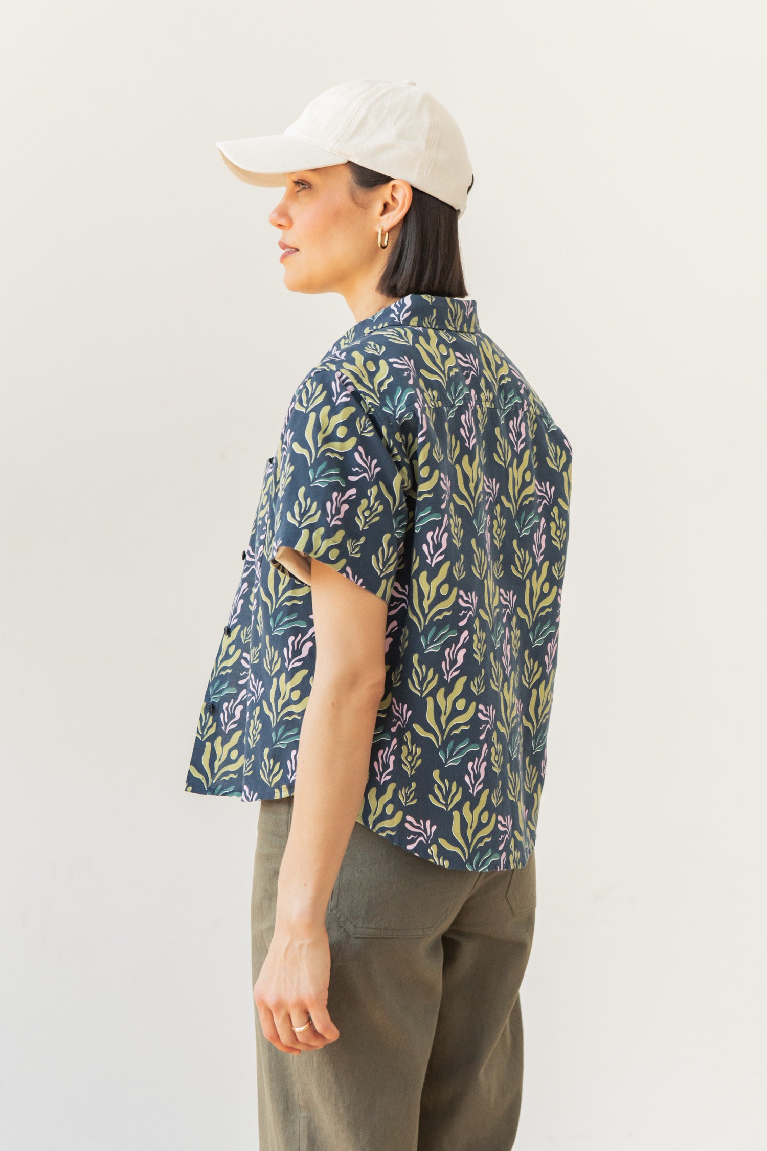 A person wearing the Bridge & Burn Greer Cropped Shirt in Spring Kelp, featuring a leafy design, paired with olive green pants and a white baseball cap, stands sideways against a plain white background.