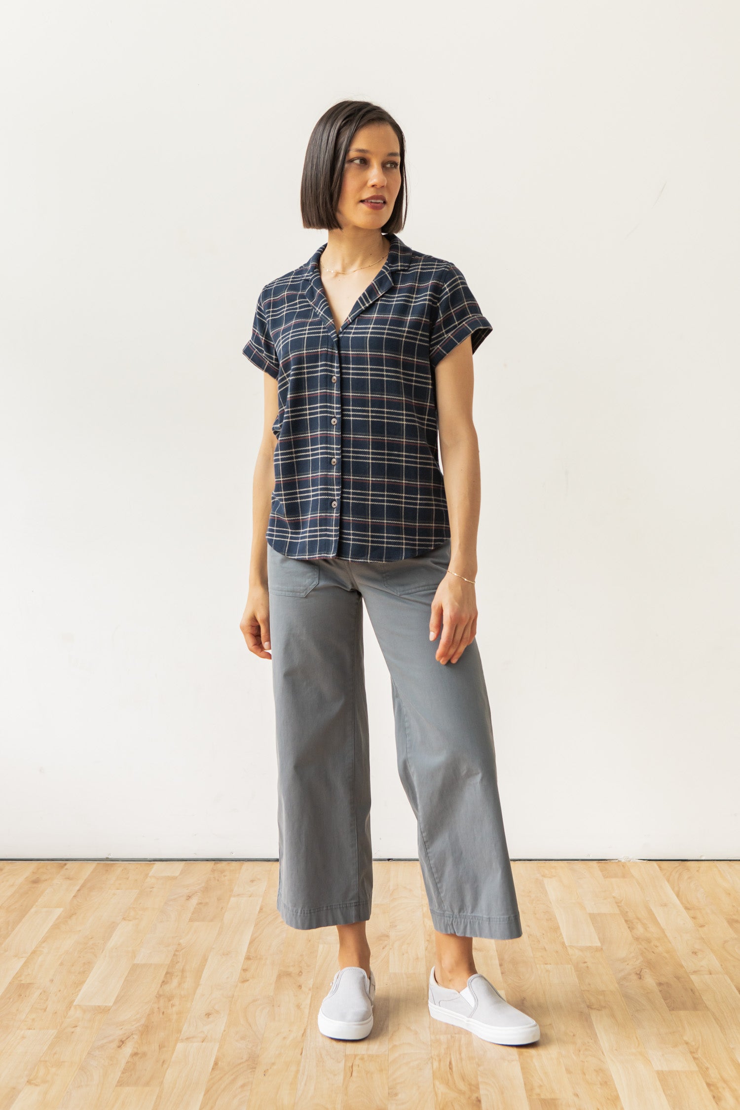 A woman stands on a wooden floor against a plain white wall, wearing the Bridge & Burn Innes Shirt in Dusk Herringbone Plaid, paired with gray wide-leg pants and white slip-on shoes. She looks slightly to her left.