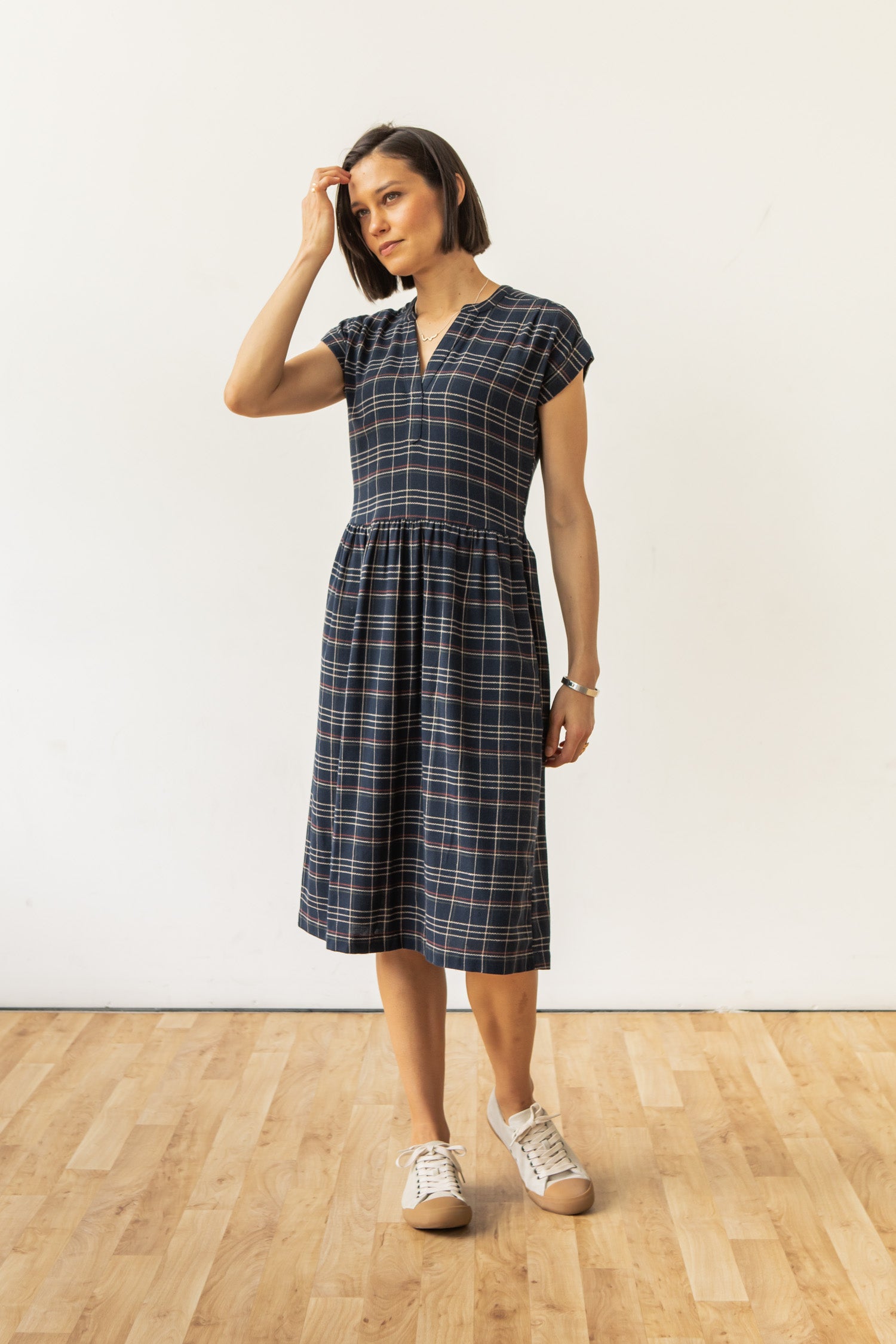The Bridge & Burn Ira Dress in Dusk Herringbone Plaid is a navy pullover with short sleeves, v-neckline, gathered waist, and beige-red plaid. Made from recycled cotton for sustainability. Shown laid flat on a white background.