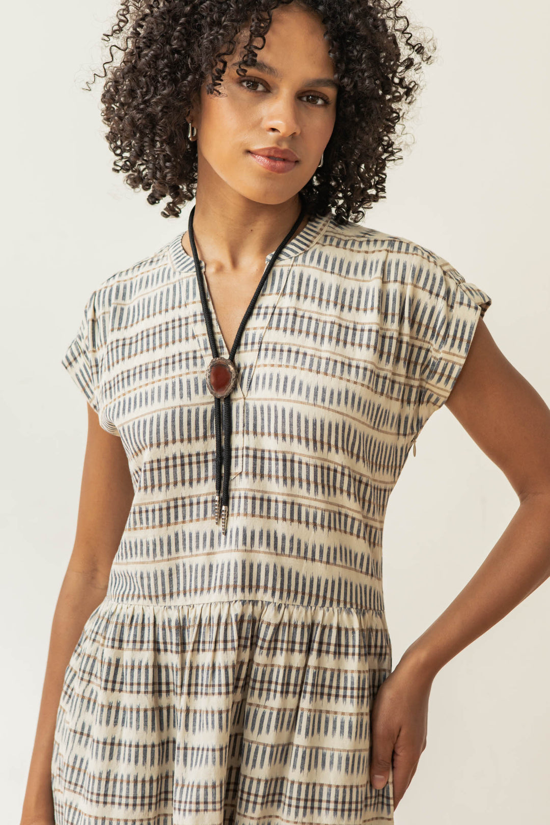 A woman with curly hair wears the Bridge & Burn Ira Dress in Mushroom Ikat Stripe, a short-sleeved sustainable dress, paired with a long necklace with a round pendant. She stands against a plain background, gazing slightly to the side.