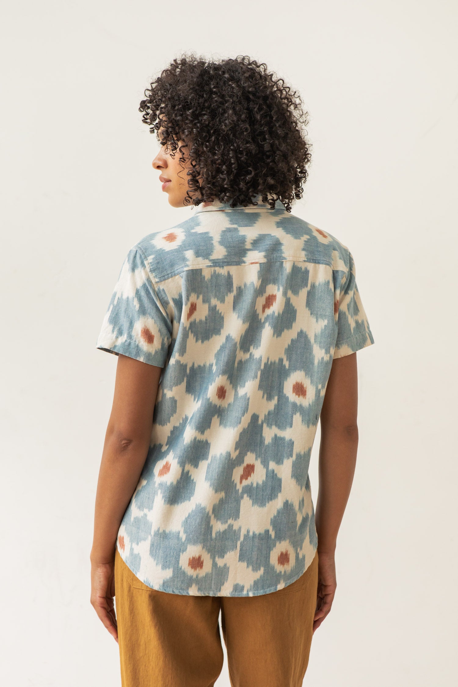 A person with short curly hair, seen from the back, wears the Bridge & Burn Lana Shirt in Polar Blossom—a relaxed fit, short sleeve button-up with a blue and beige abstract pattern—paired with brown pants against a plain light background.