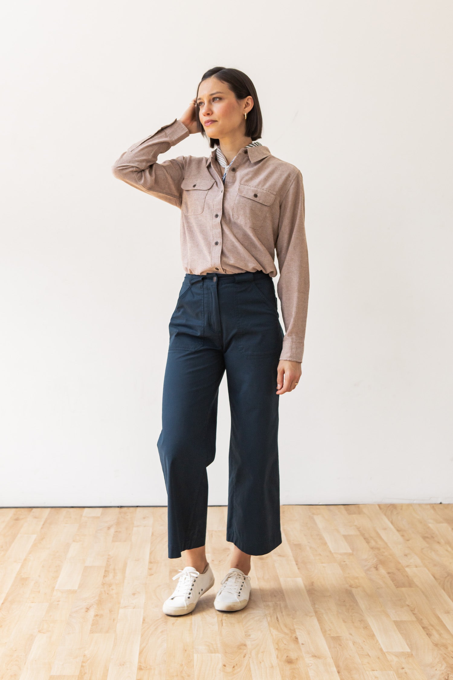 A woman stands indoors on a wooden floor wearing the Bridge & Burn Lenon Shirt in Rust Chambray, navy high-waisted wide-leg pants, and white sneakers, with one hand touching her head as she looks off to the side.