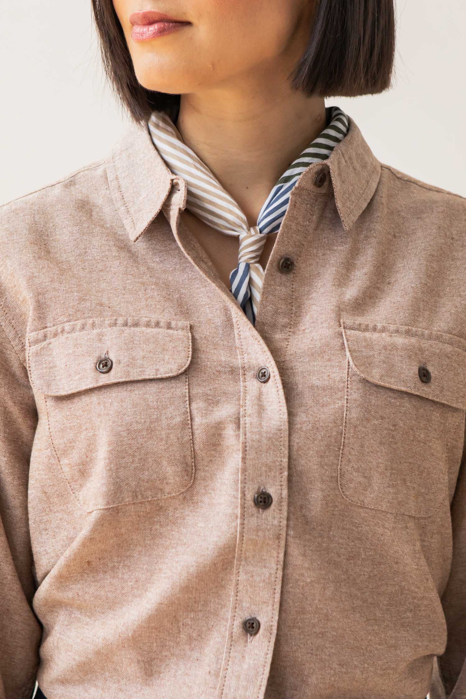 A woman wears the Bridge & Burn Lenon Shirt in Rust Chambray, a relaxed fit style with chest pockets made from recycled cotton, paired with a striped neck scarf. Only her lower face and upper body are visible.