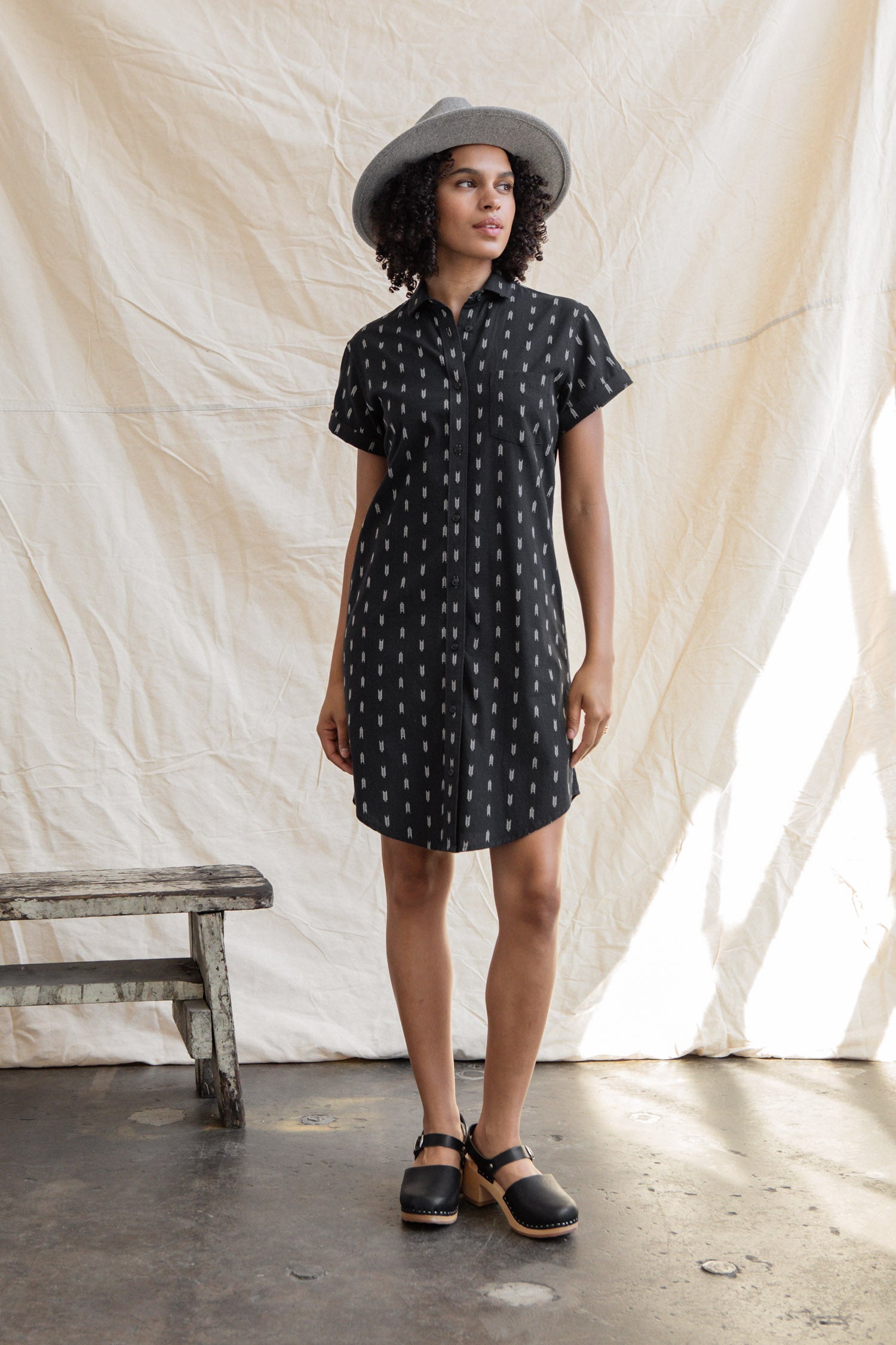 The Loren Shirt Dress in Onyx Arrows by Bridge & Burn is a short-sleeved, knee-length black cotton dress with a collar, subtle white vertical pattern, button front, and curved hem—ideal for versatile workwear.