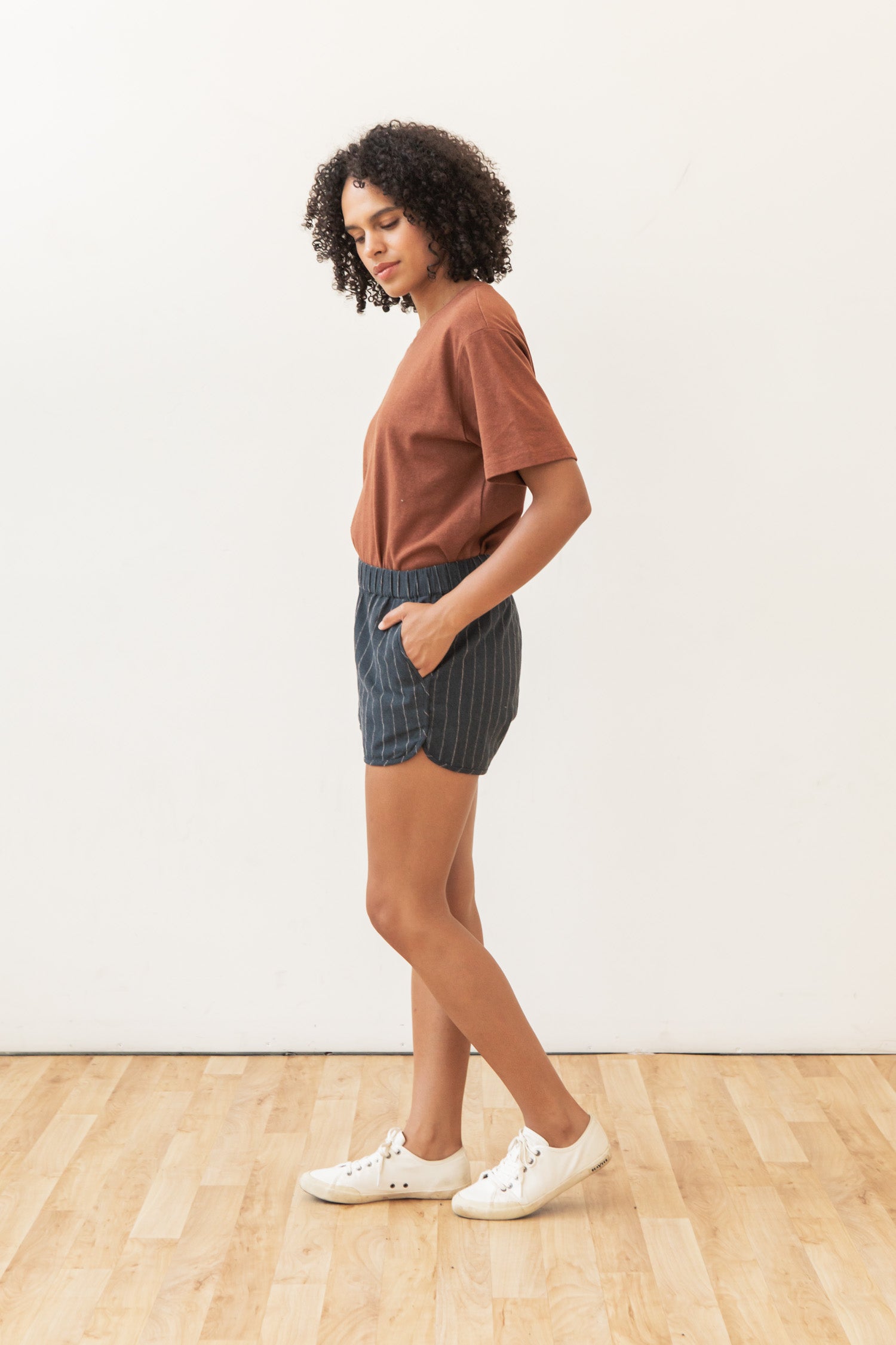 A woman with curly hair stands sideways on a wooden floor, wearing a brown t-shirt, Bridge & Burn's Luca Track Short in Charcoal Stripe, and white sneakers, with her hands in her pockets against a plain white background.