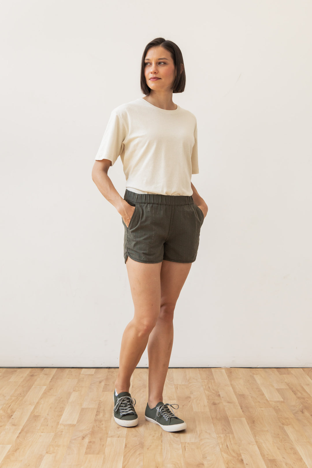 A woman stands on a wooden floor, wearing a short-sleeve cream t-shirt and Bridge & Burn’s Luca Track Short in Olive Stripe with pockets, paired with gray sneakers. She faces sideways with her hands in her pockets against a white wall.