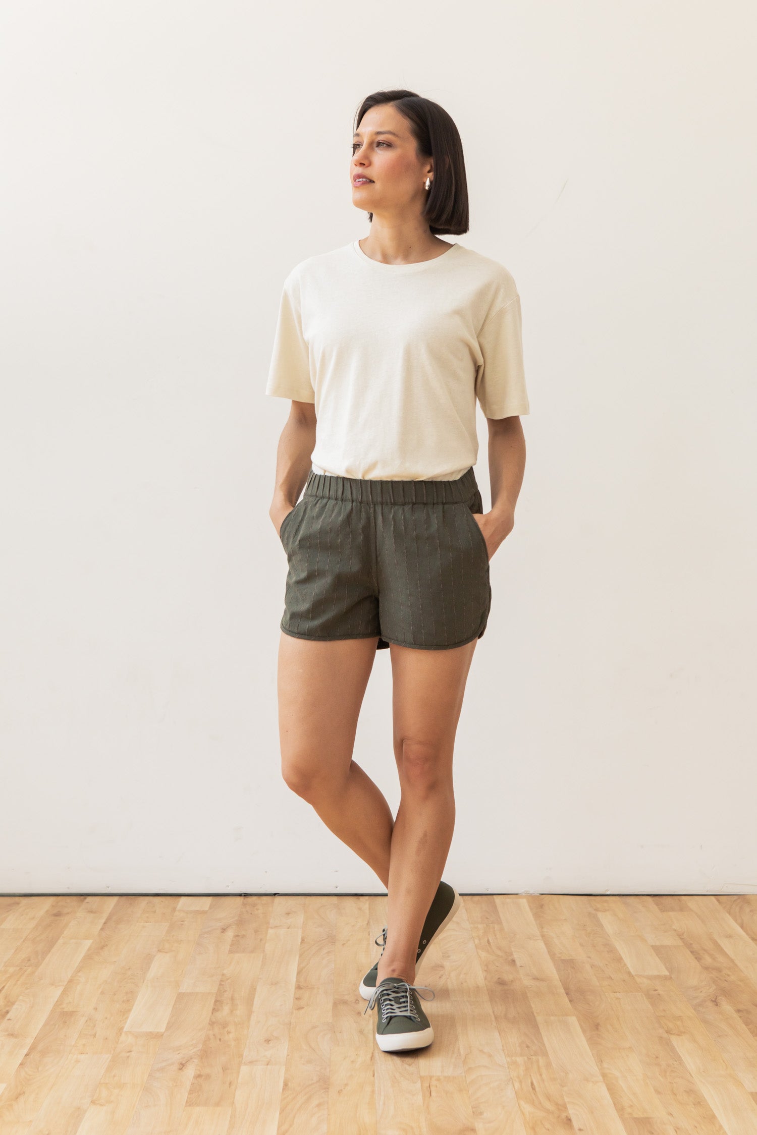 A woman with short dark hair stands on a wooden floor by a white wall, wearing a white t-shirt, green sneakers, and Bridge & Burn's Luca Track Short in Olive Stripe, looking to the side with her hands in her pockets.