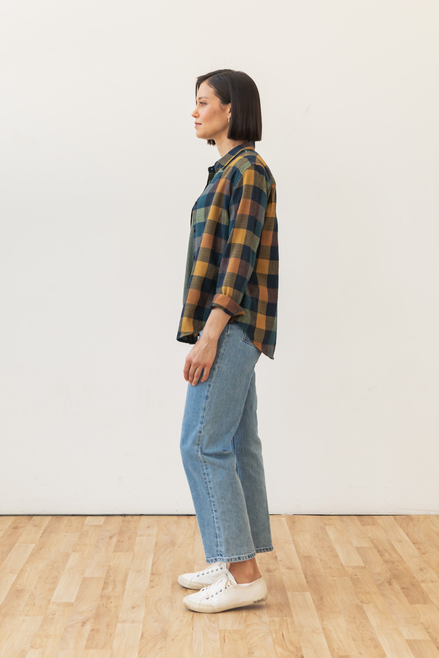 A person with short dark hair stands in profile on a wooden floor, wearing the Bridge & Burn Olivia Slim Shirt in Moonbow Plaid, light blue jeans, and white shoes, against a plain off-white wall.