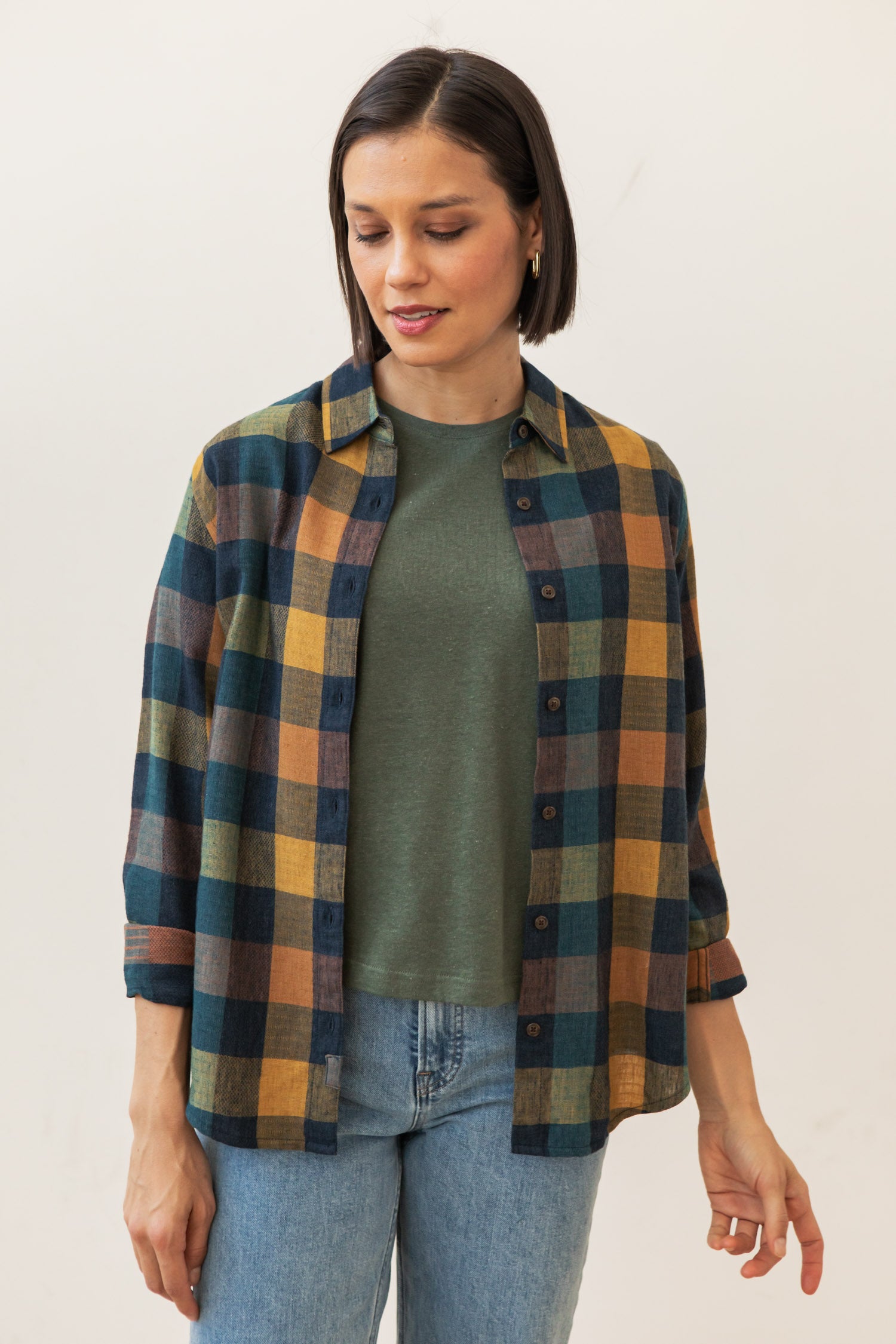 A woman with short dark hair wears a green tee, light blue jeans, and the Bridge & Burn Olivia Slim Shirt in Moonbow Plaid. She looks down with a slight smile against a plain light background.
