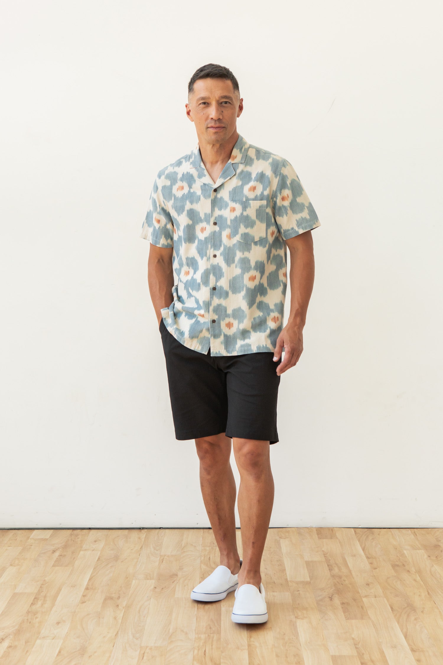 A man stands indoors on a wooden floor in front of a white wall, wearing the Bridge & Burn Bodie Shirt in Polar Blossom, black shorts, and white slip-on shoes, with one hand in his pocket.