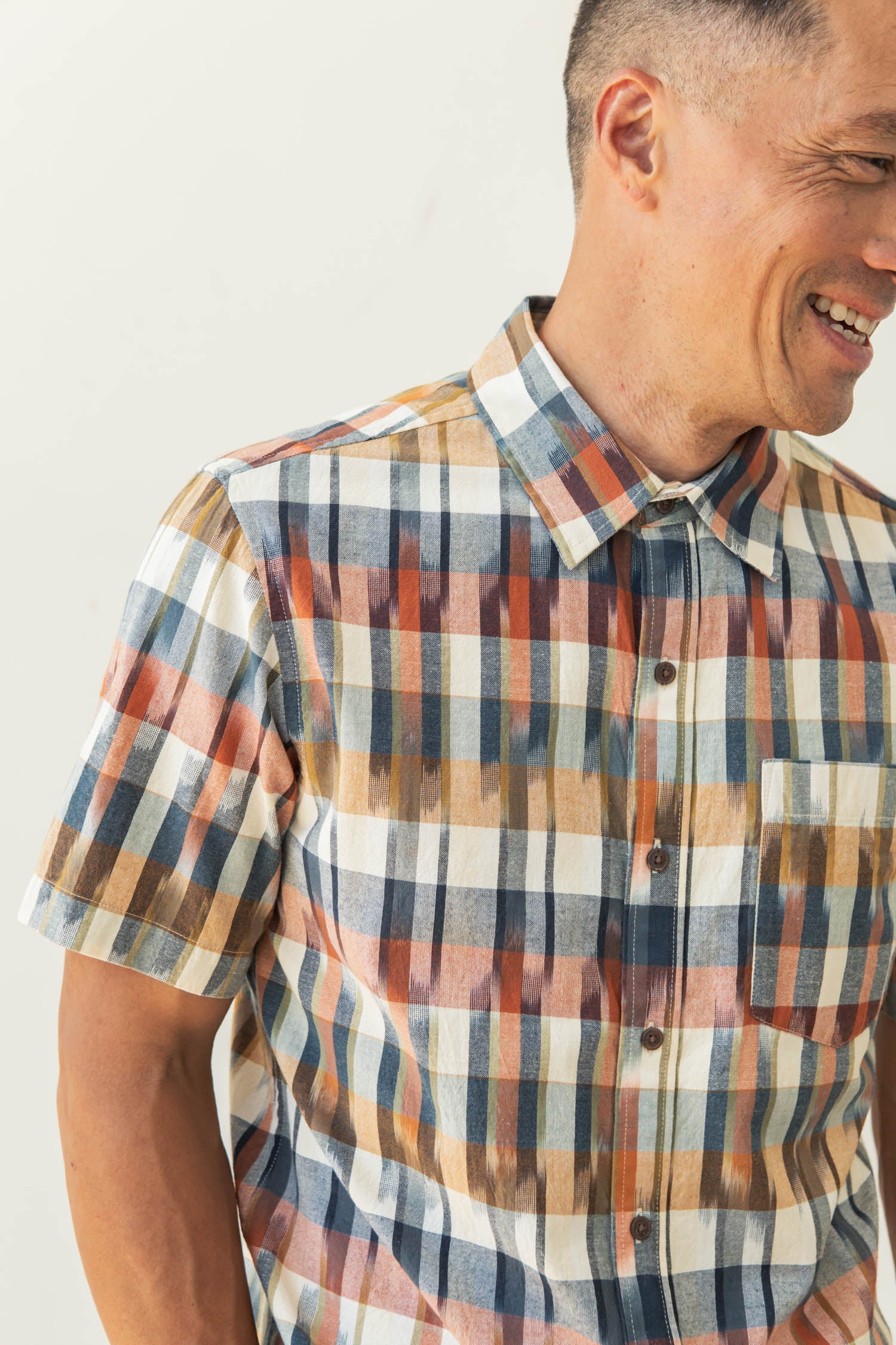 A man smiles wearing the Bridge & Burn Harbor Slim Shirt in Aurora Space Dye Plaid, a short-sleeved button-up with a colorful blue, orange, yellow, and cream plaid pattern. He faces slightly right with only part of his face visible.