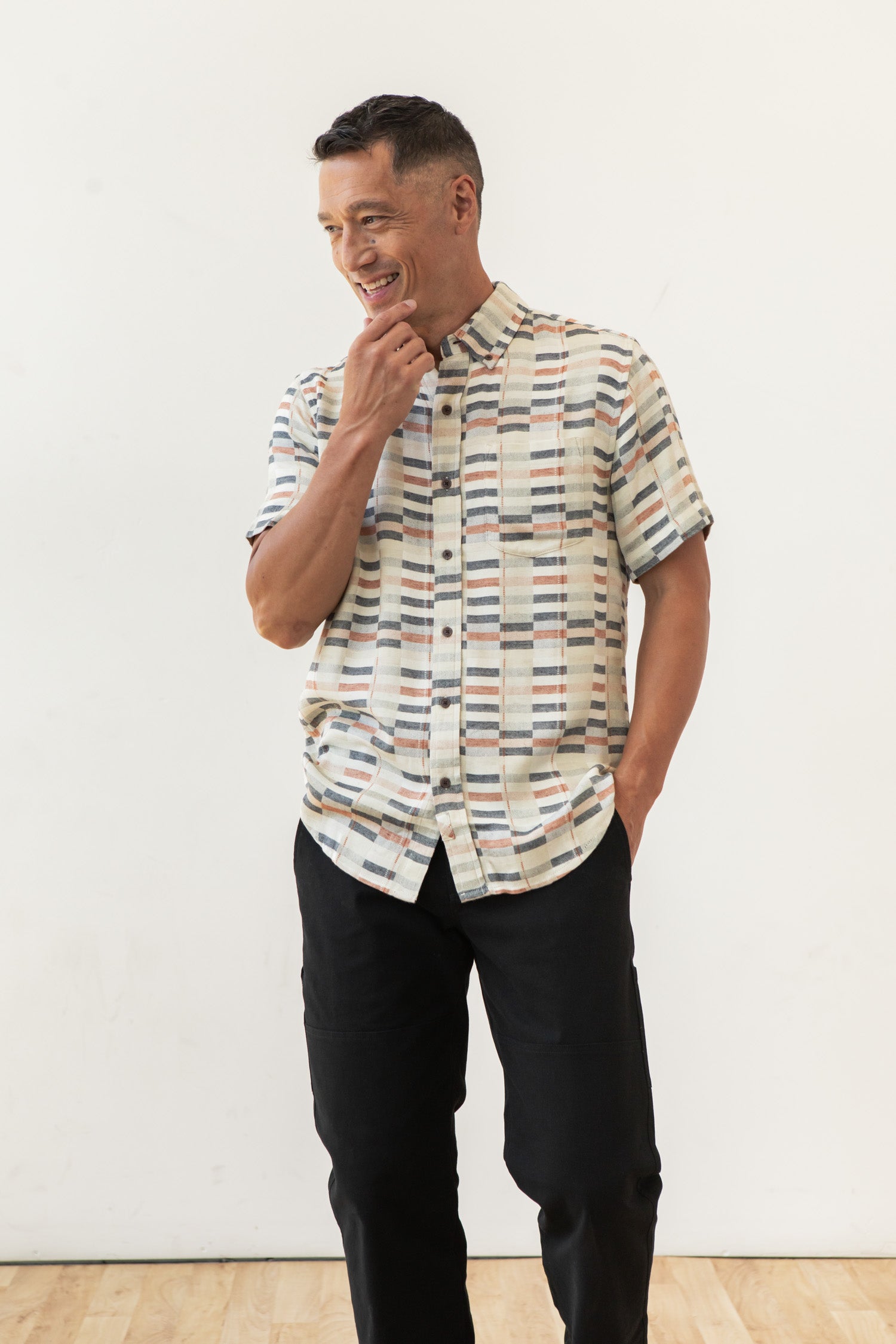 A man smiles against a white background, wearing the Bridge & Burn Jordan Slim Shirt in Broadway Stripe with black pants. One hand is in his pocket, the other touches his chin—showcasing this modern wardrobe essential.