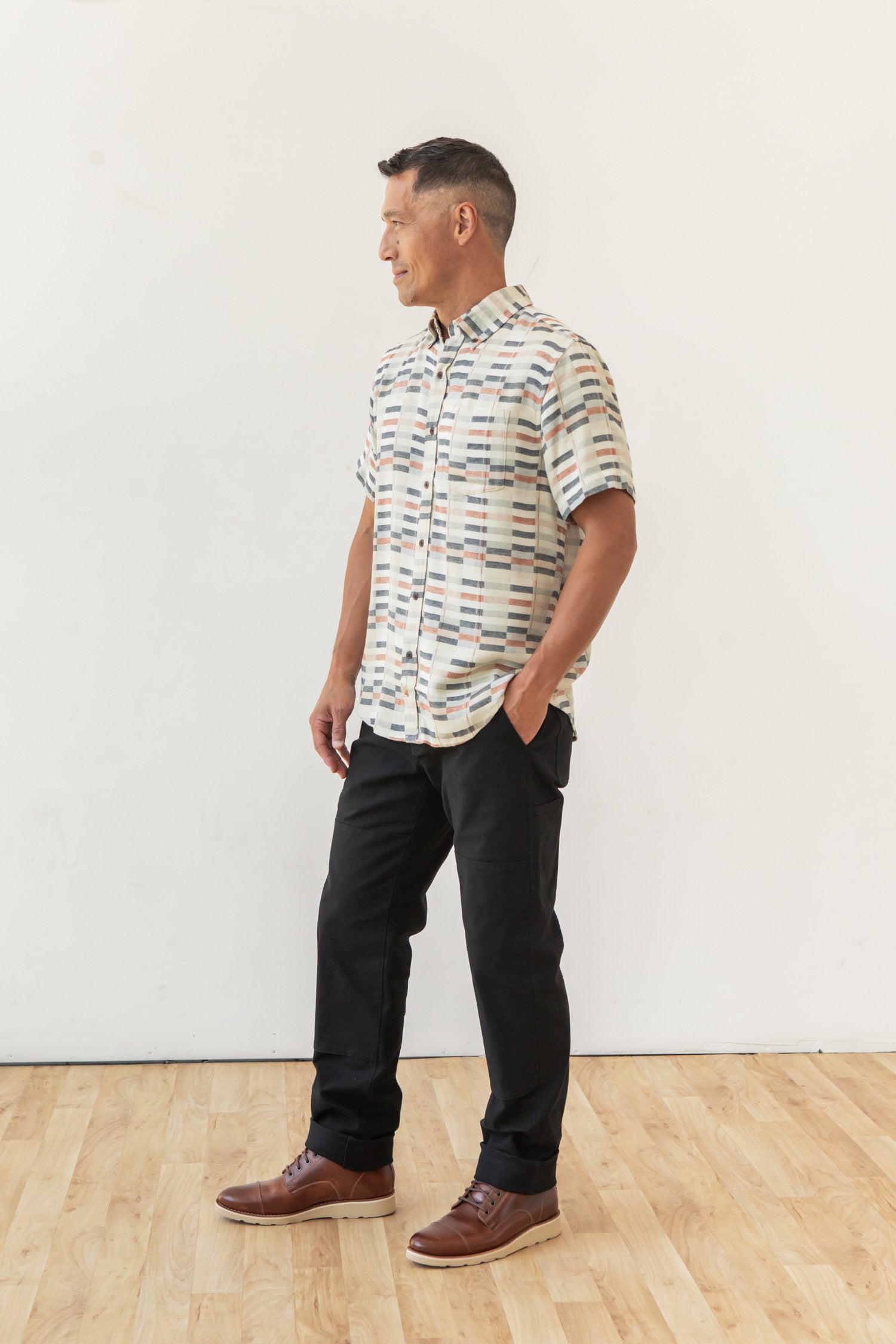 A man stands on a wooden floor against a white wall, wearing the Bridge & Burn Jordan Slim Shirt in Broadway Stripe with black pants and brown shoes—hands in his pockets, looking aside. This shirt is a key piece for contemporary style.