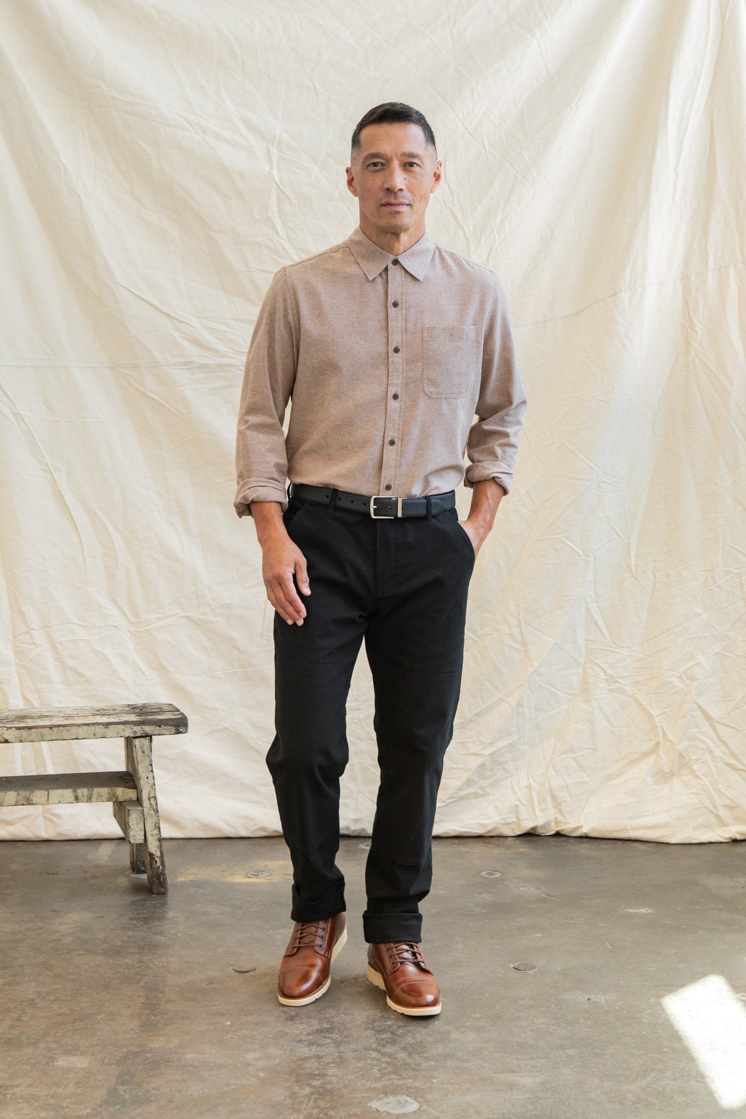 A man stands indoors before a cream fabric backdrop, wearing the Bridge & Burn Larson Shirt in rust chambray, black pants with a black belt, and brown shoes. One hand in his pocket, he stands next to a wooden bench.