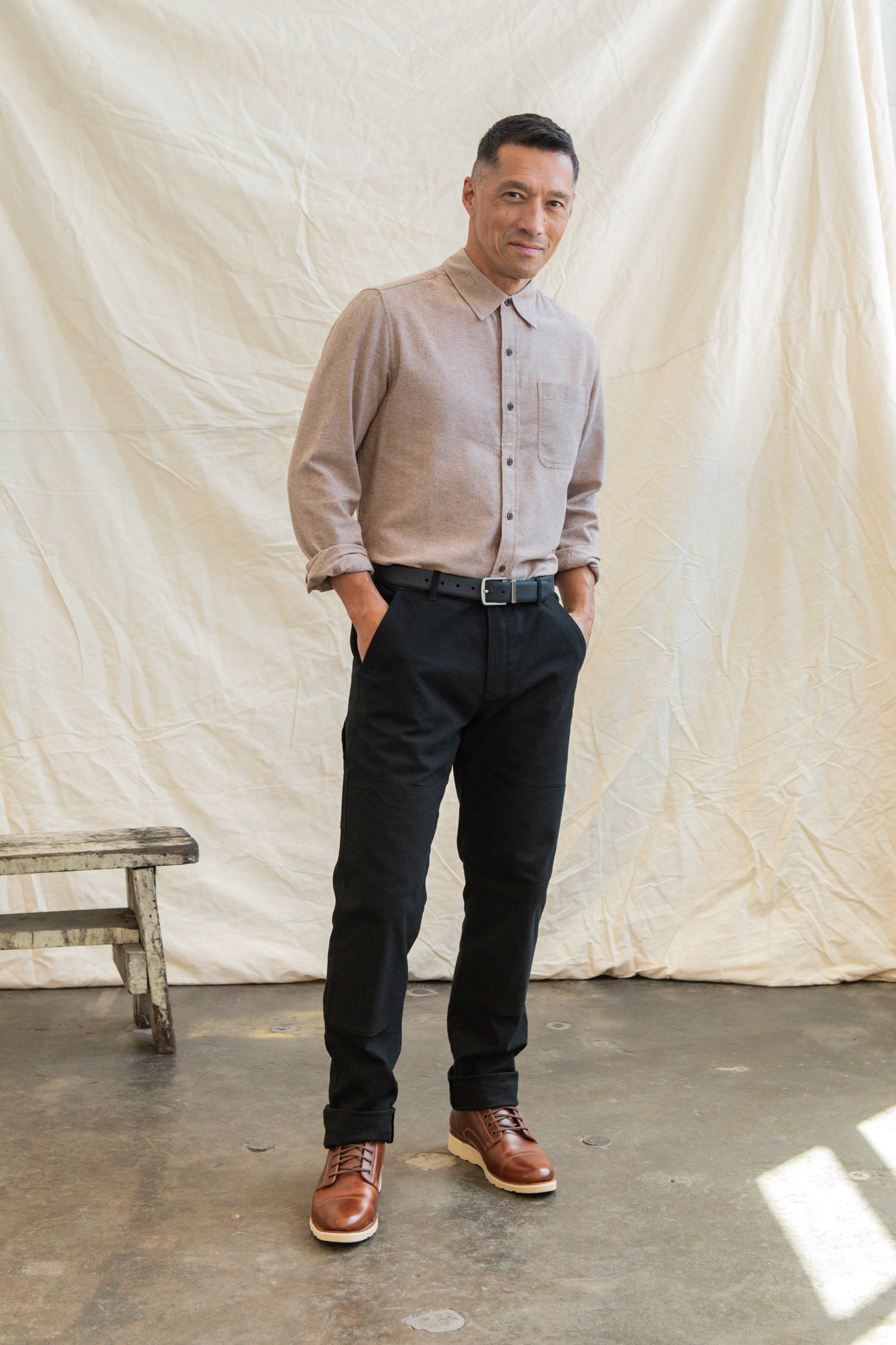 A man stands indoors with his hands in his pockets, wearing the Bridge & Burn Larson Shirt in Rust Chambray, black pants, and brown boots. A white fabric backdrop and wooden stool are visible behind him.