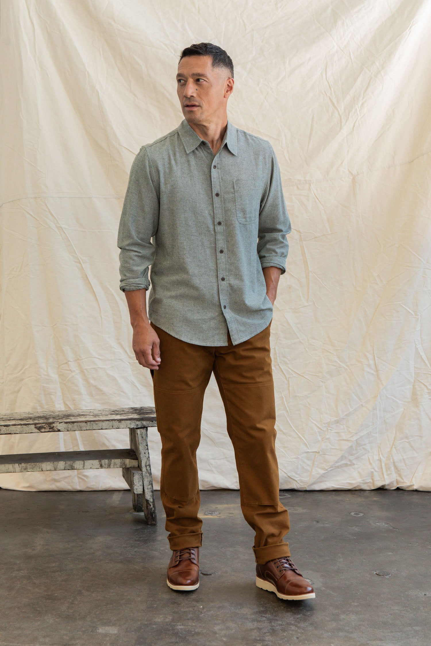 A man stands indoors on a concrete floor, wearing the Bridge & Burn Larson Shirt in Sage Chambray, brown pants, and brown shoes. He looks to the side with one hand in his pocket; a wooden bench and beige backdrop are behind him.